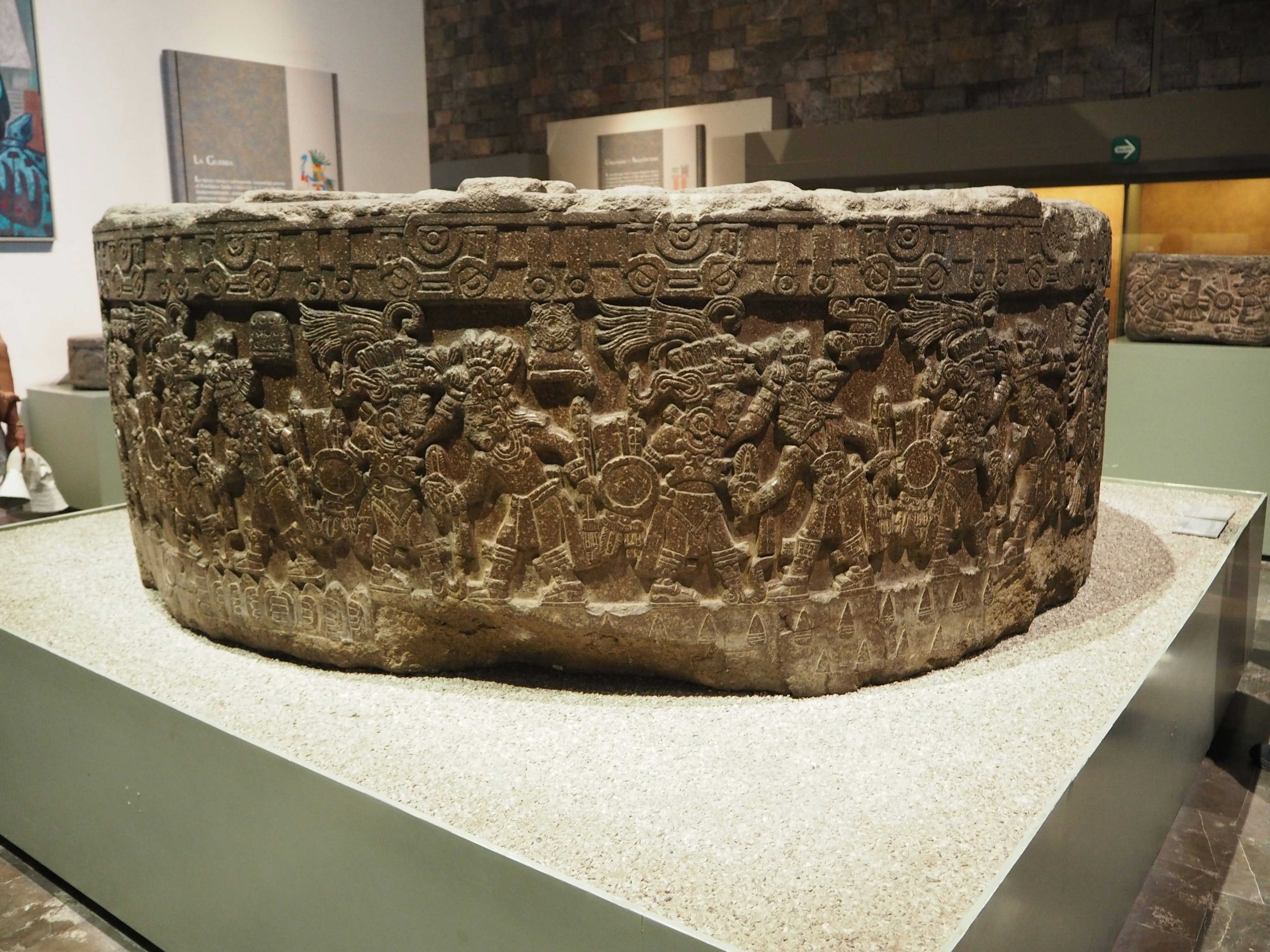 Round stone sculpture displayed in a museum with various human figures carved on its surface