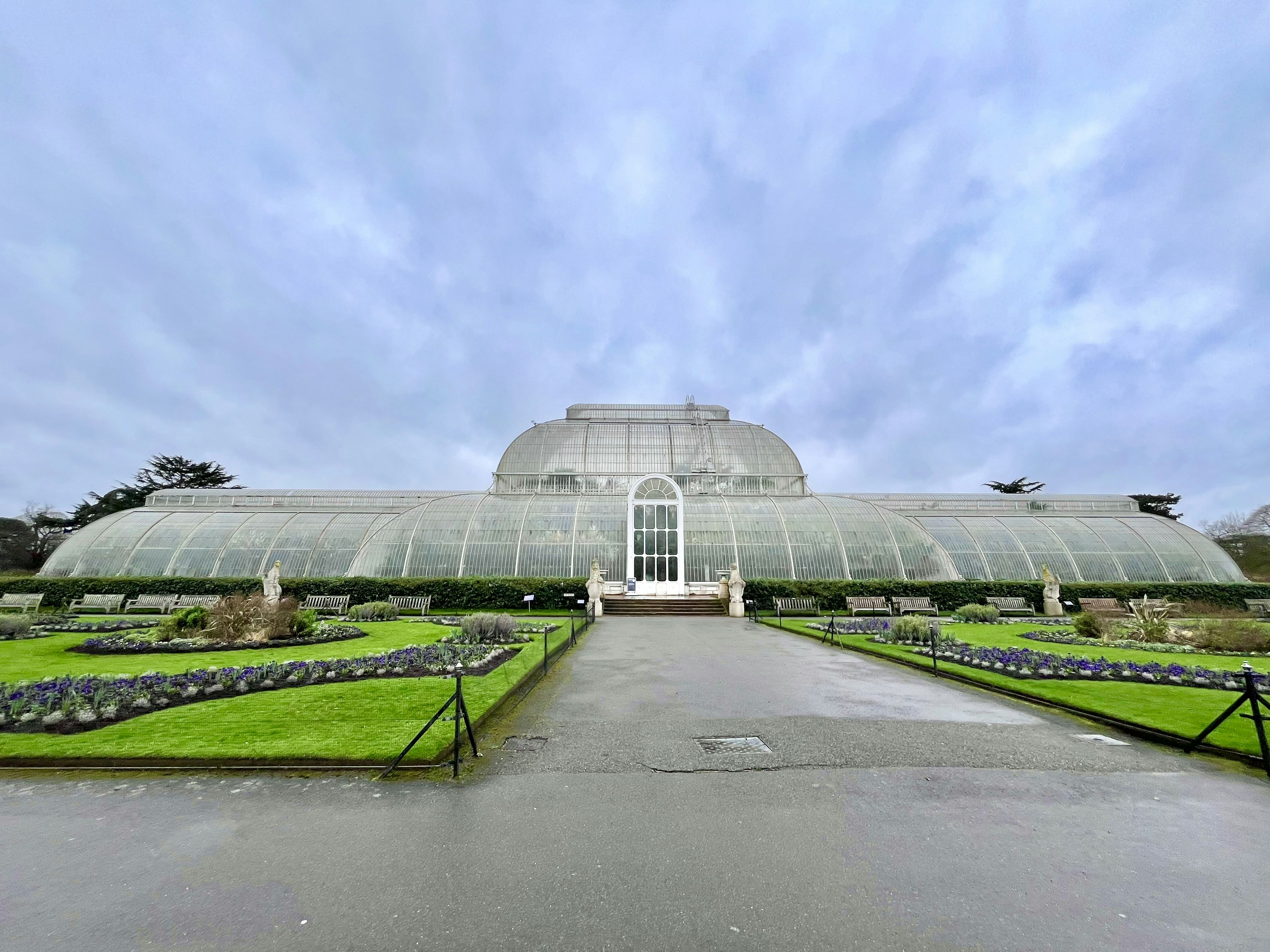 Vista frontal de un gran invernadero de vidrio rodeado de jardines verdes y cielo nublado