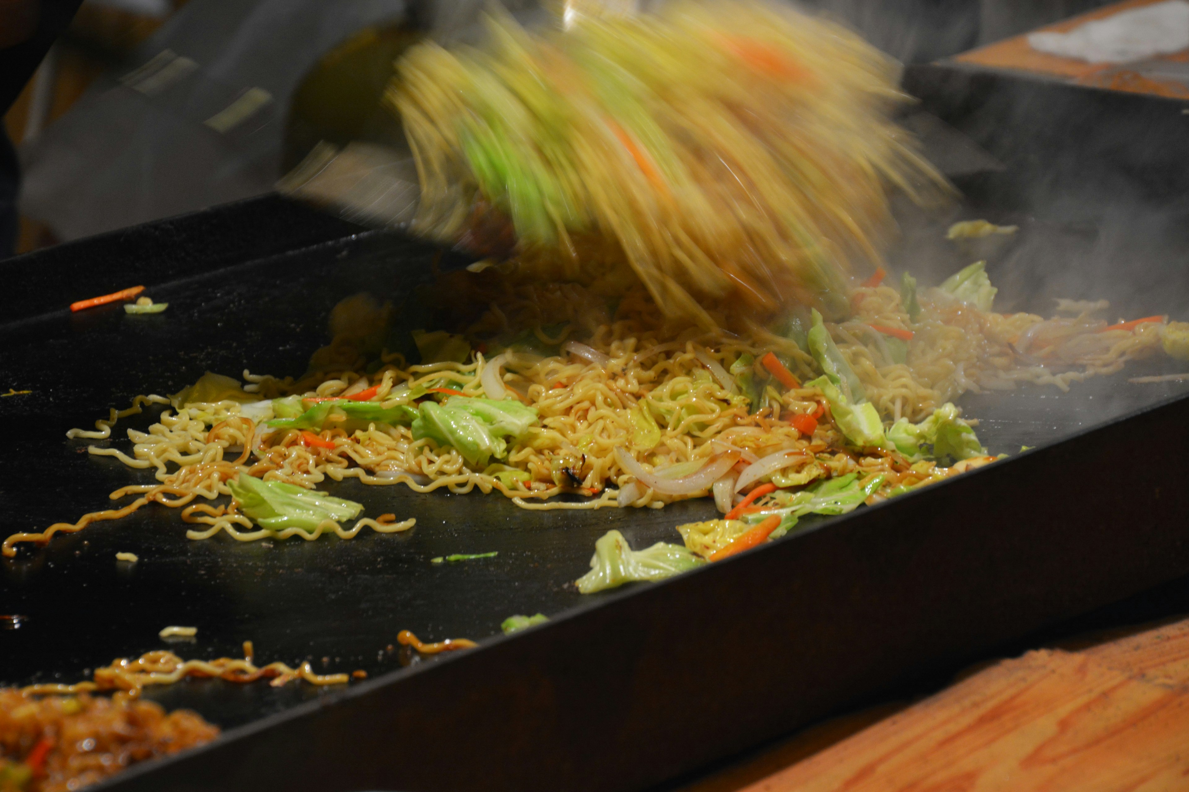 Stir-fried noodles and vegetables being tossed on a hot griddle