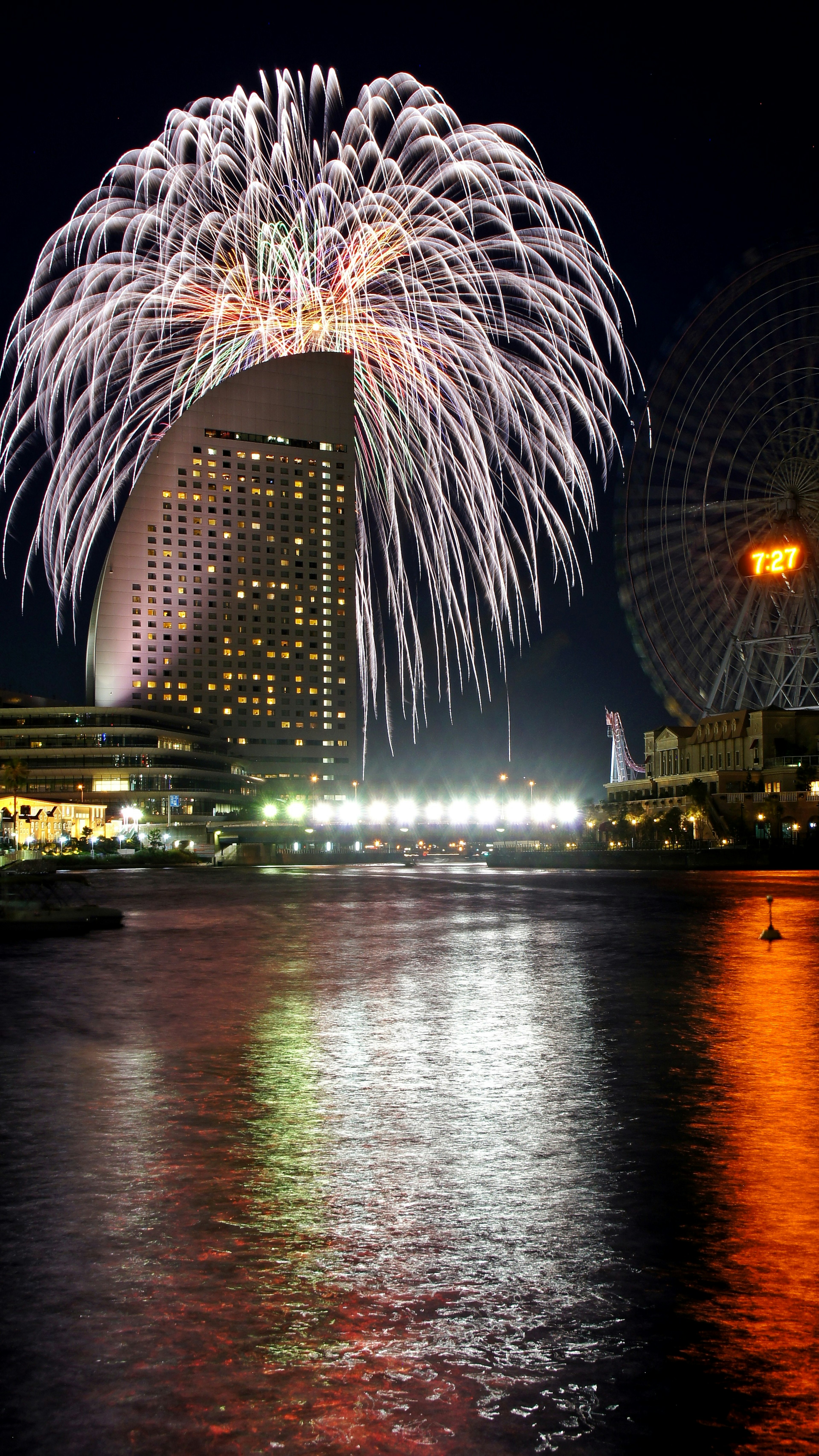 Fireworks display over a river with illuminated buildings at night