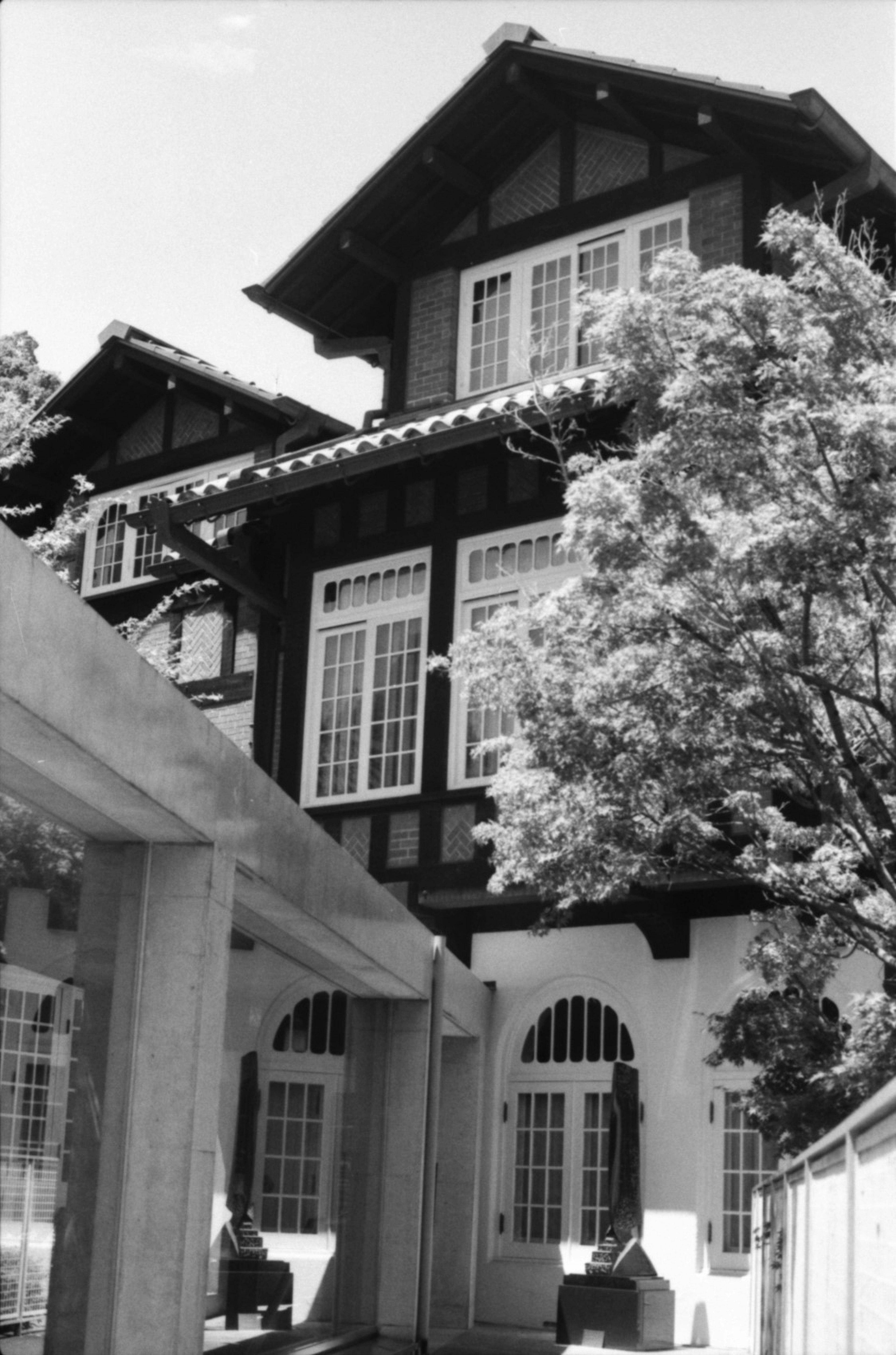 Black and white image of a historic building exterior featuring distinctive wooden roof and large windows surrounded by green trees