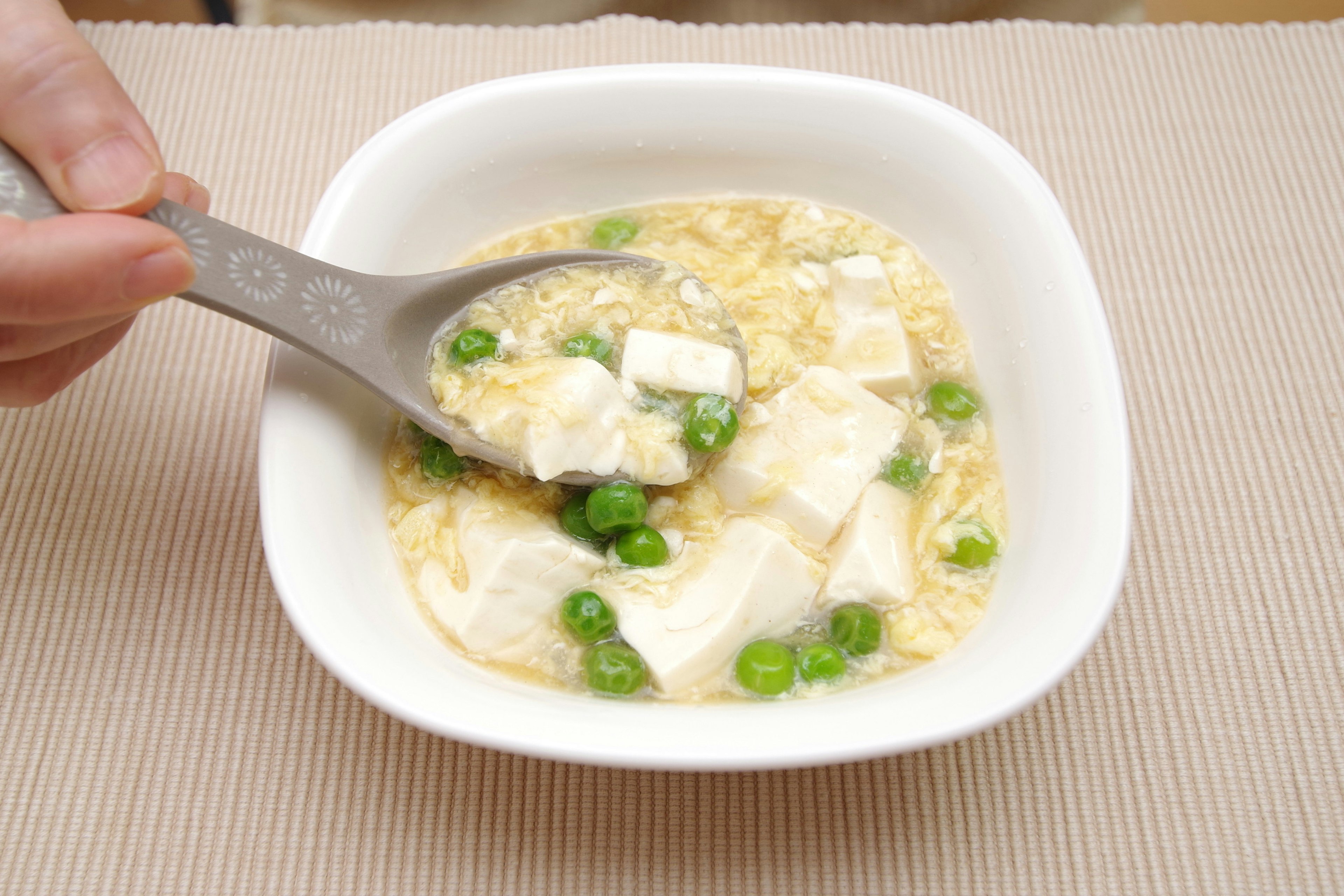 Una mano mezclando sopa de tofu y guisantes verdes con una cuchara