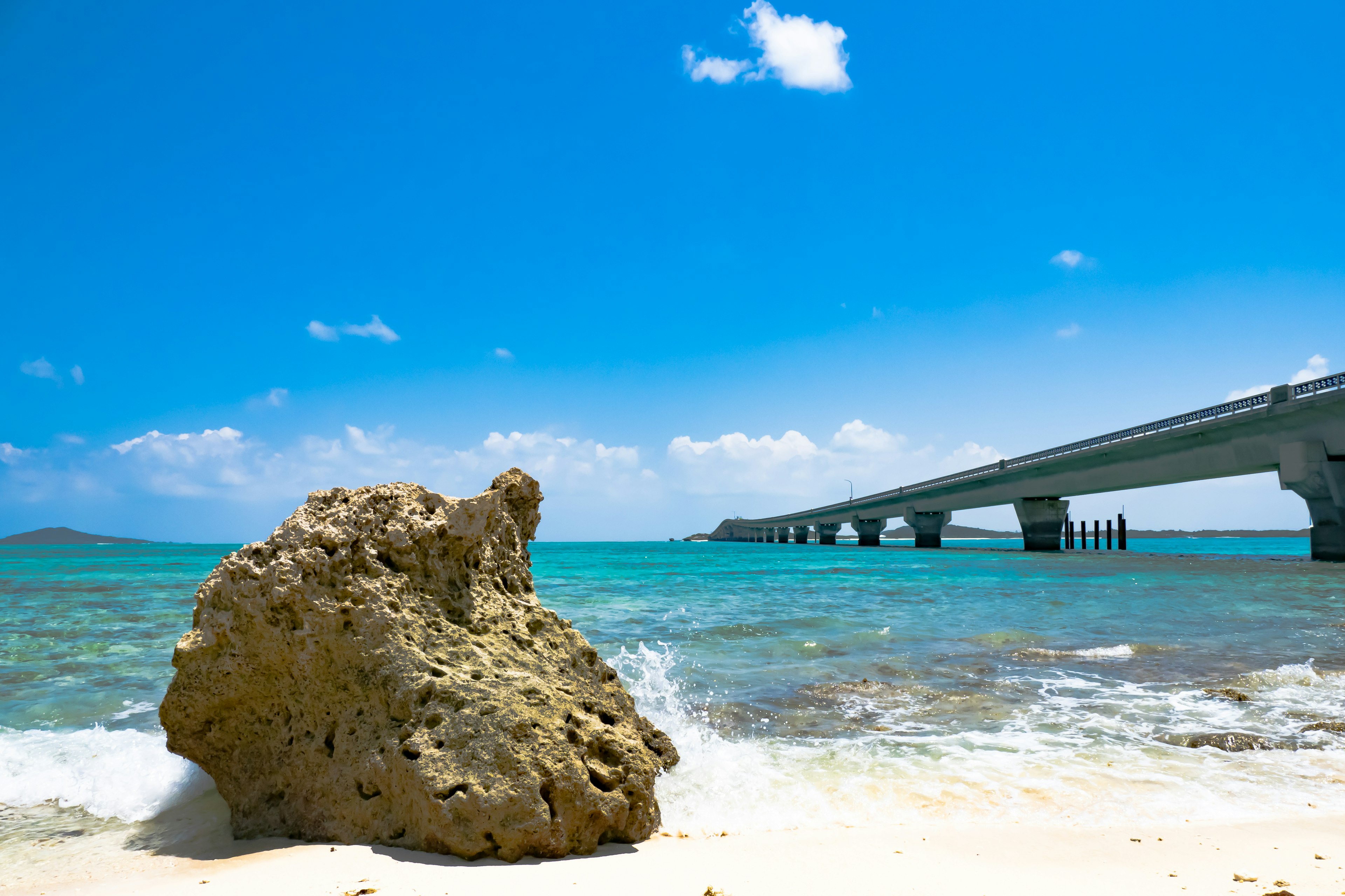 Una gran roca en una playa con agua azul clara y cielo, un puente al fondo