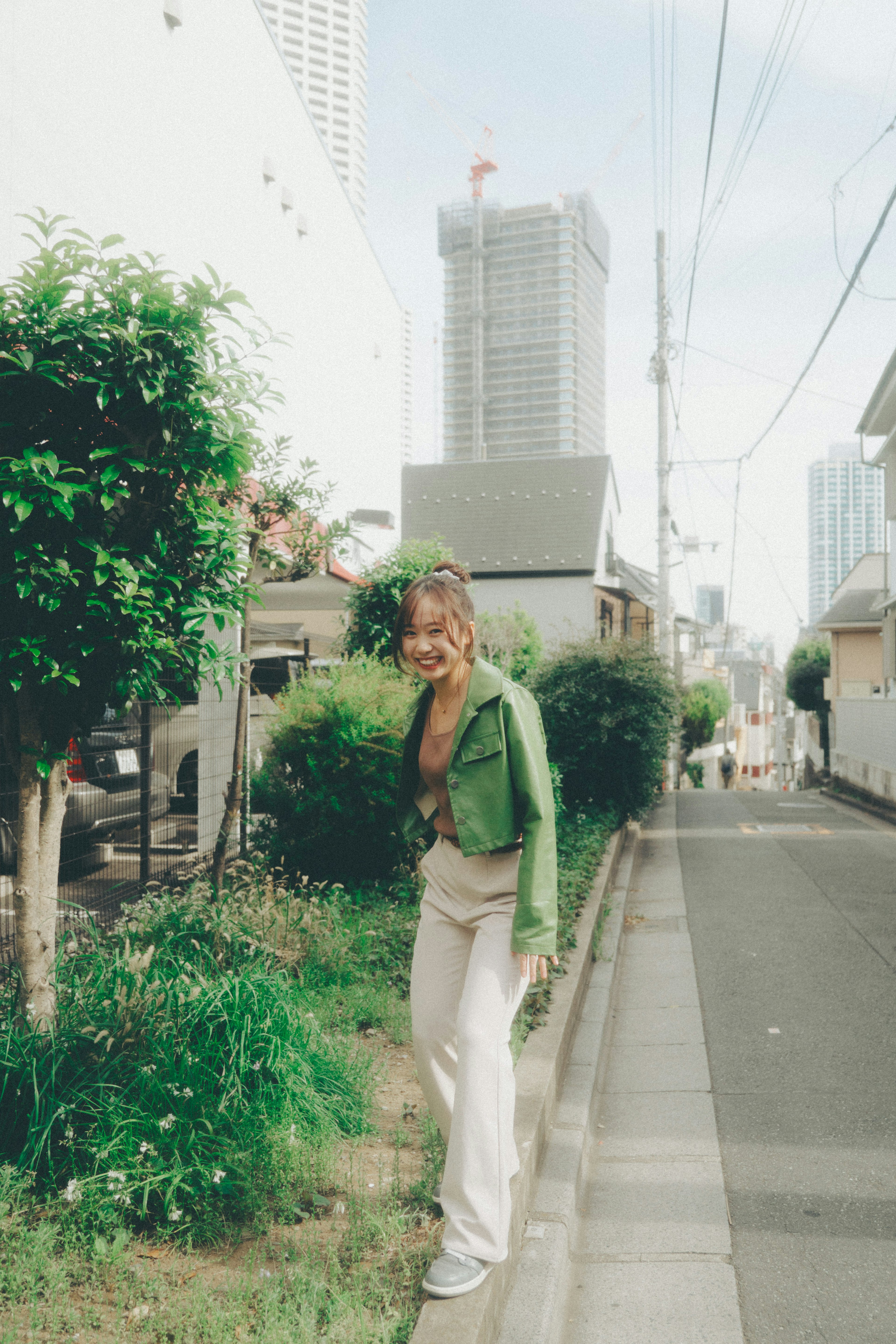 A woman in a green jacket stands on a street with tall buildings and greenery in the background