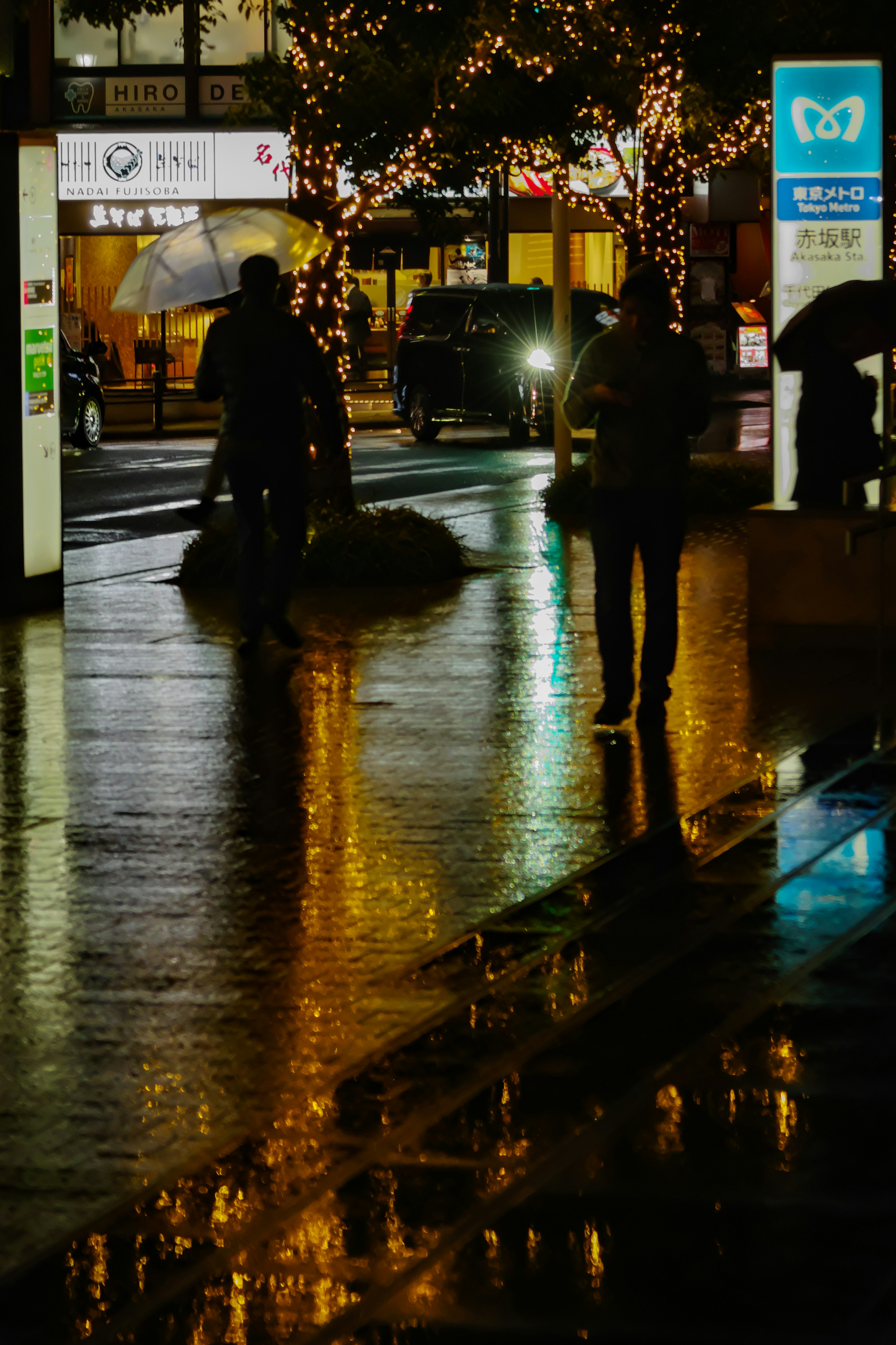 夜間城市景觀中，撐著傘的人們在濕滑人行道上的輪廓