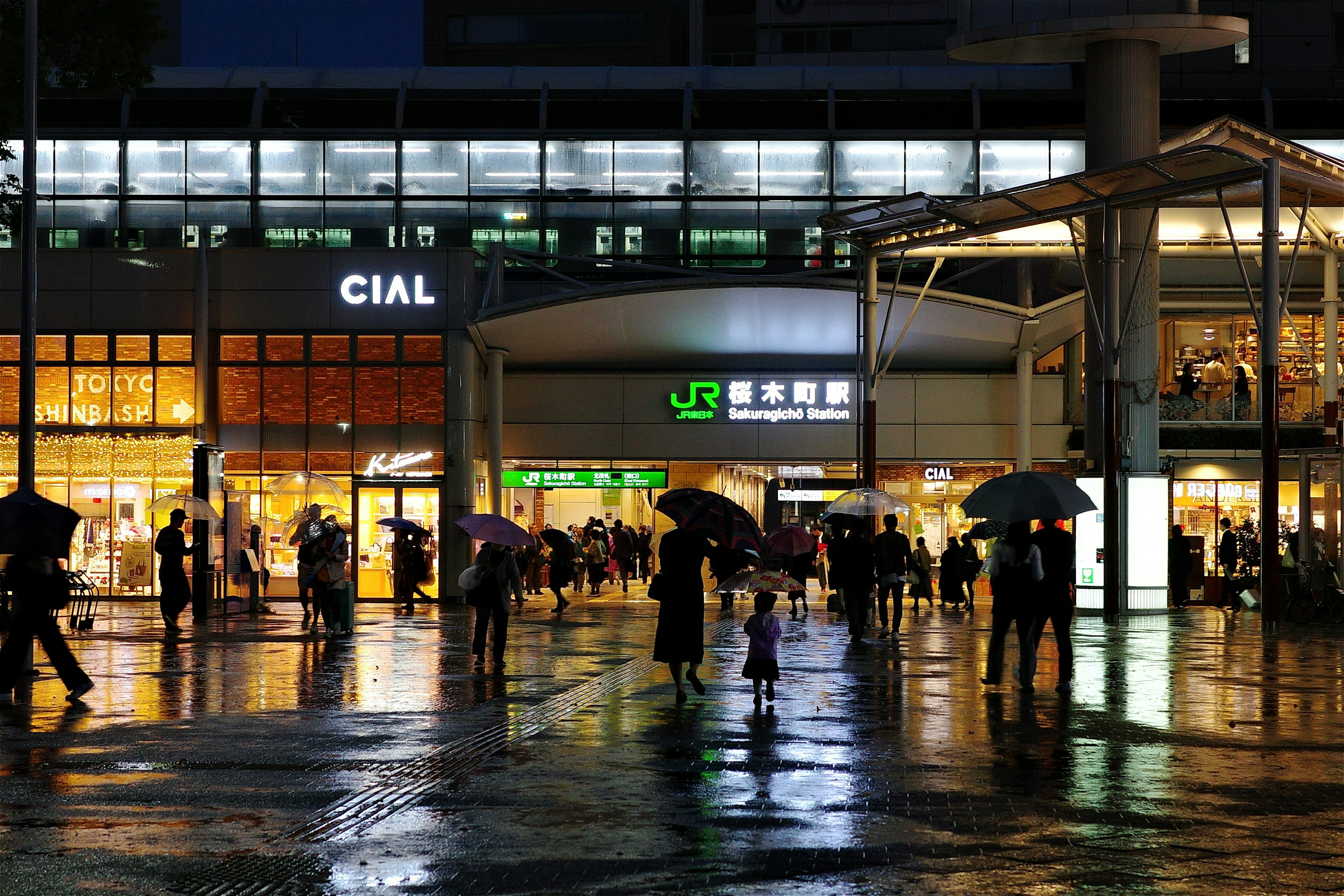 夜の駅前の風景 雨の中で傘をさした人々が歩いている 駅の看板が明るく照らされている