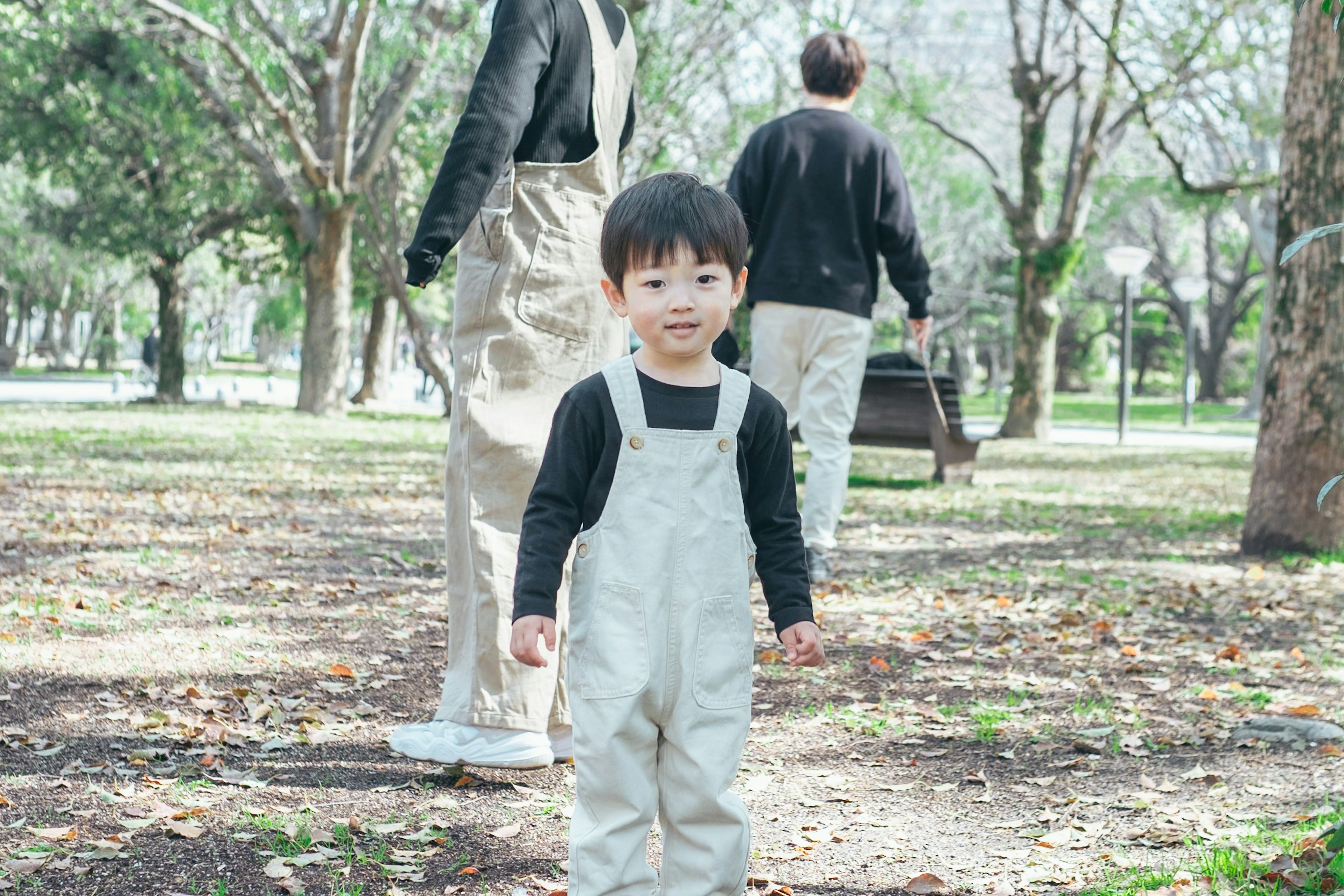 A young boy in white overalls walking in a park with two adults in the background