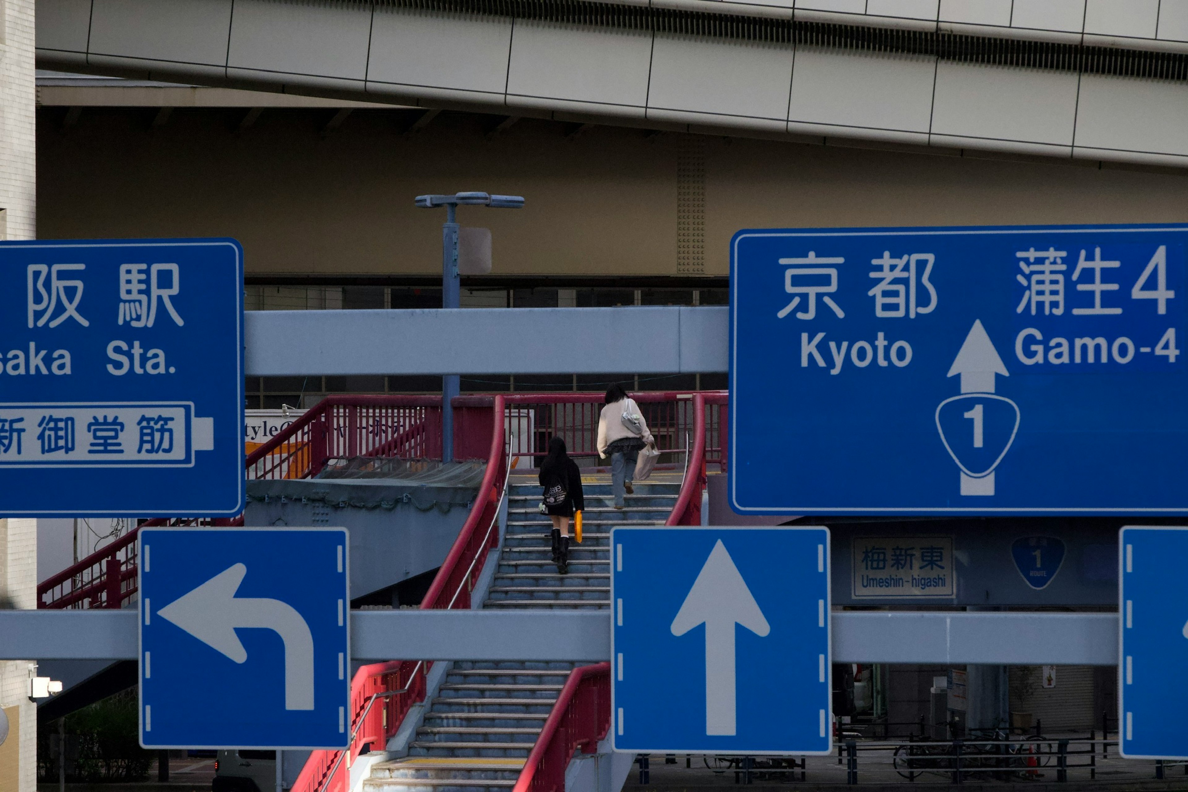 Orang-orang menaiki tangga dengan tanda biru yang menunjukkan Stasiun Osaka dan tujuan Kyoto