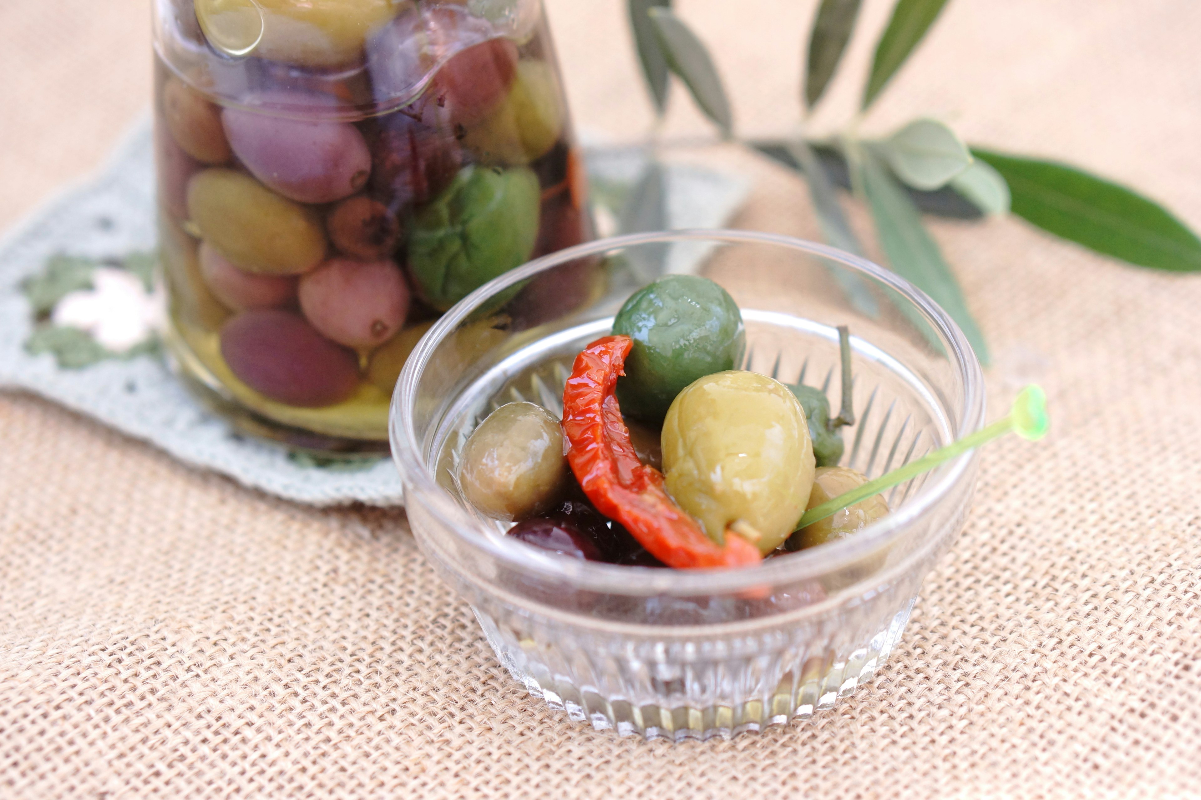 Image of a small bowl of olives and chili peppers with a jar in the background