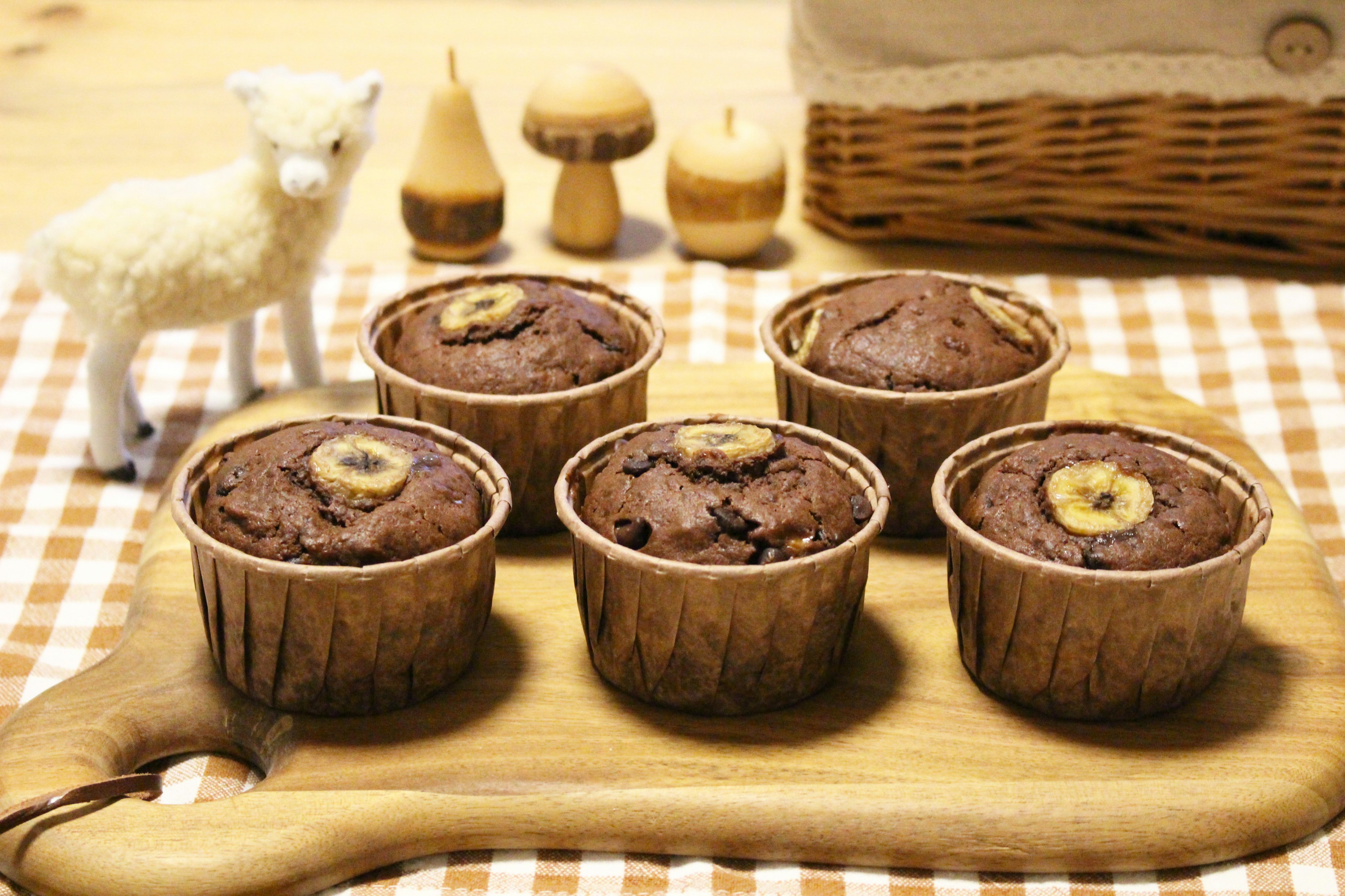 Five chocolate muffins topped with banana slices on a wooden board with a small sheep figurine and wooden objects in the background