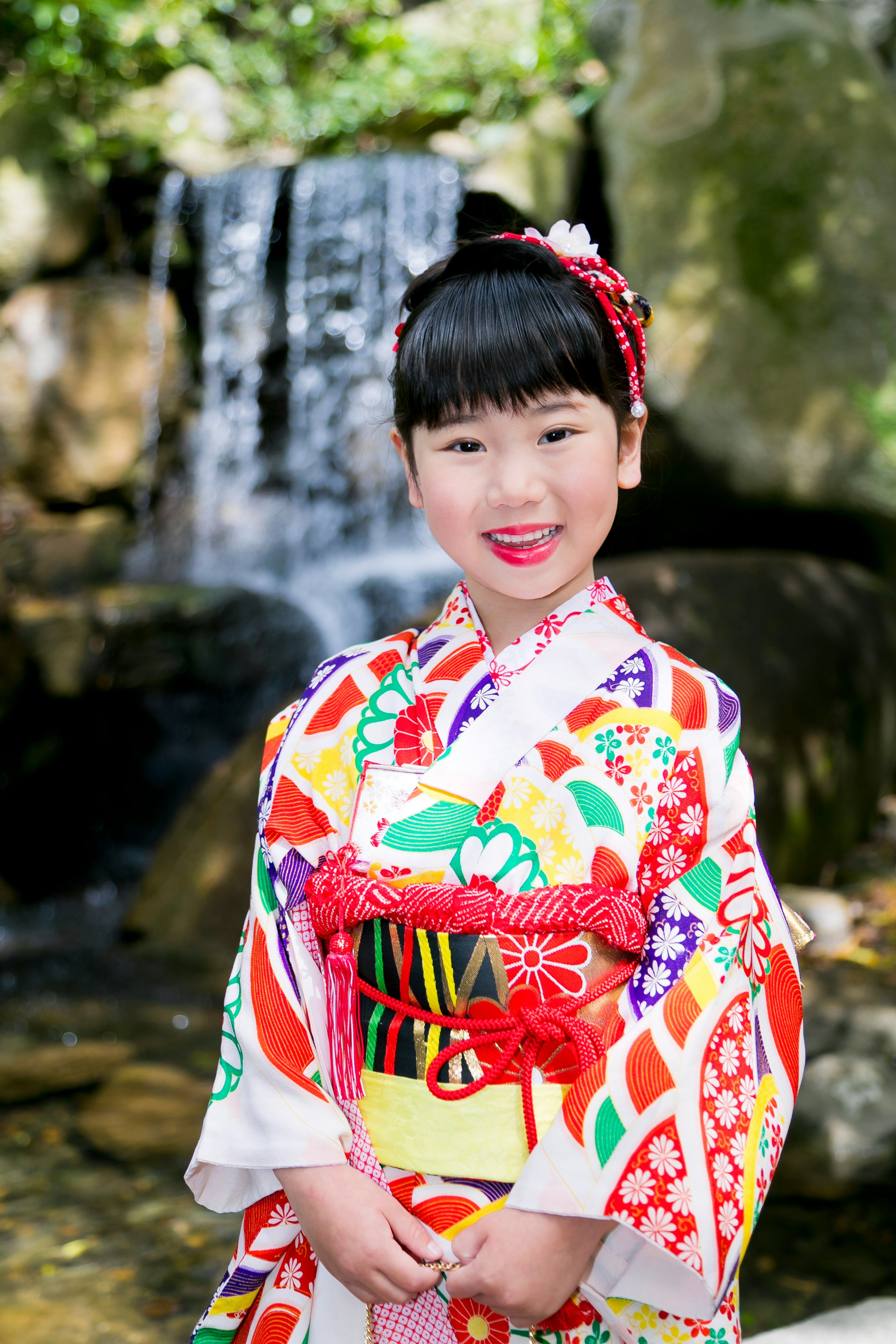 Seorang gadis dalam kimono berwarna cerah tersenyum di depan air terjun