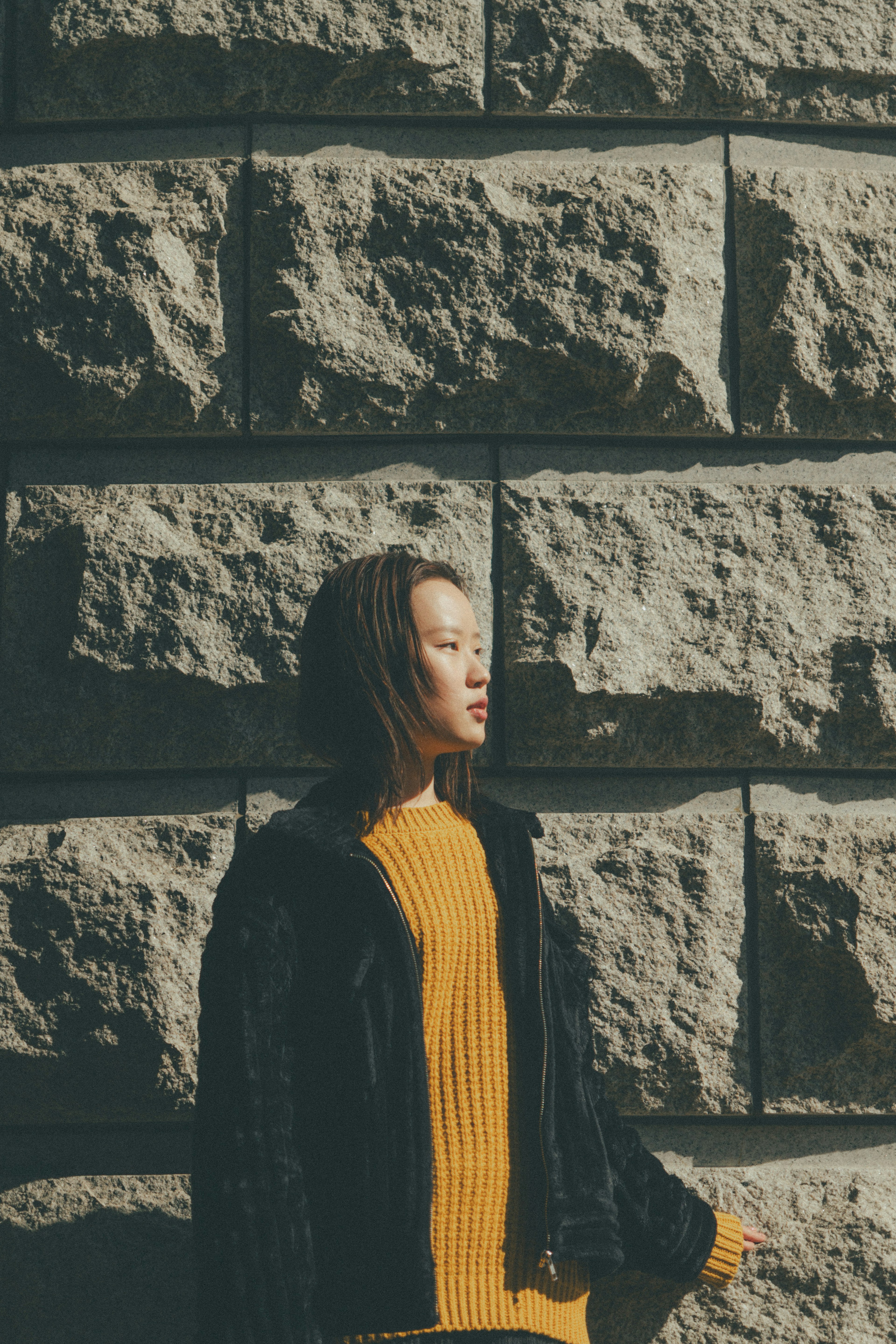 Seitenansicht einer Frau, die vor einer Steinmauer steht, trägt einen gelben Pullover und eine schwarze Jacke