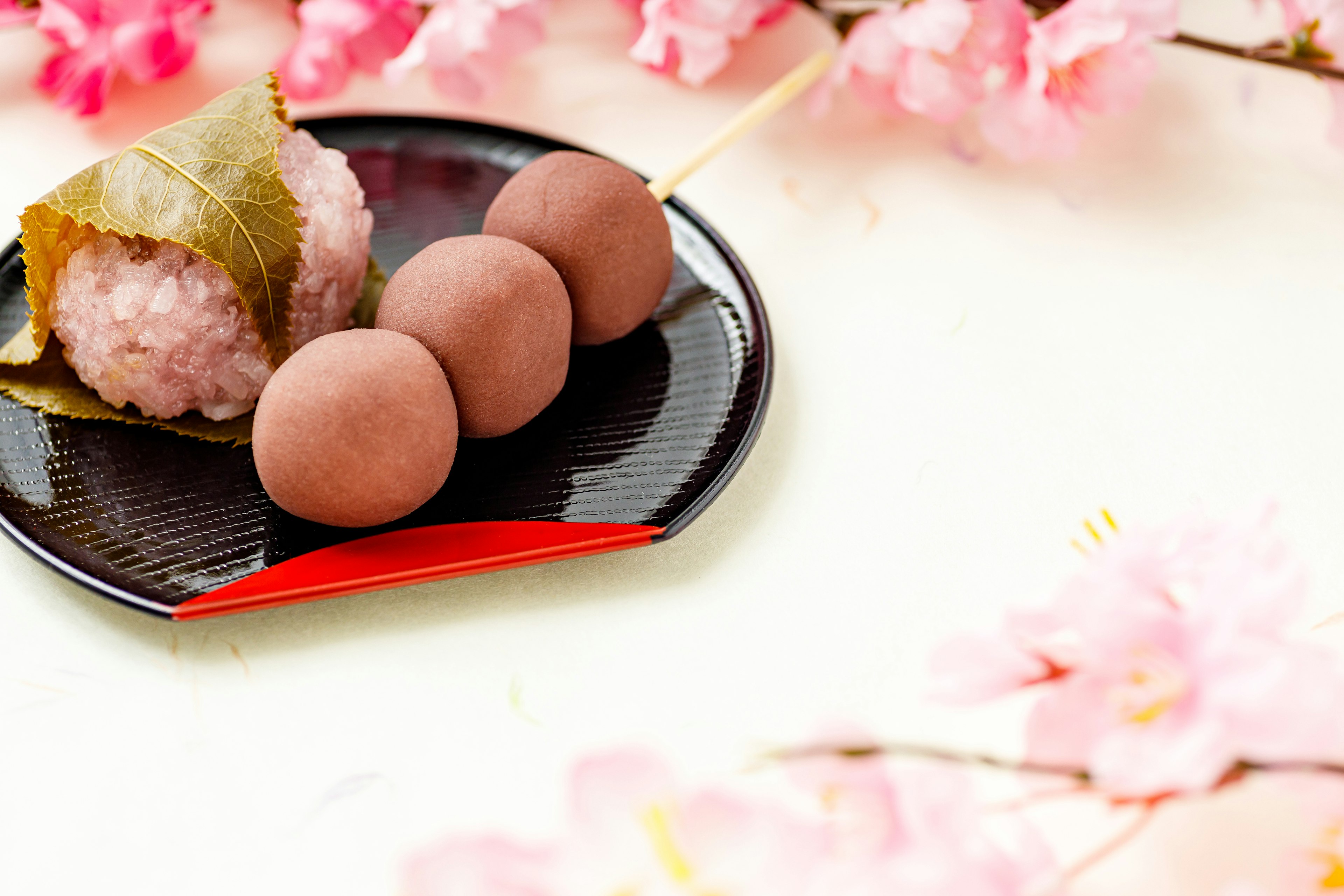 Piring kue tradisional Jepang dengan kelopak sakura di latar belakang menampilkan bola nasi merah muda dan kue nasi yang dibungkus