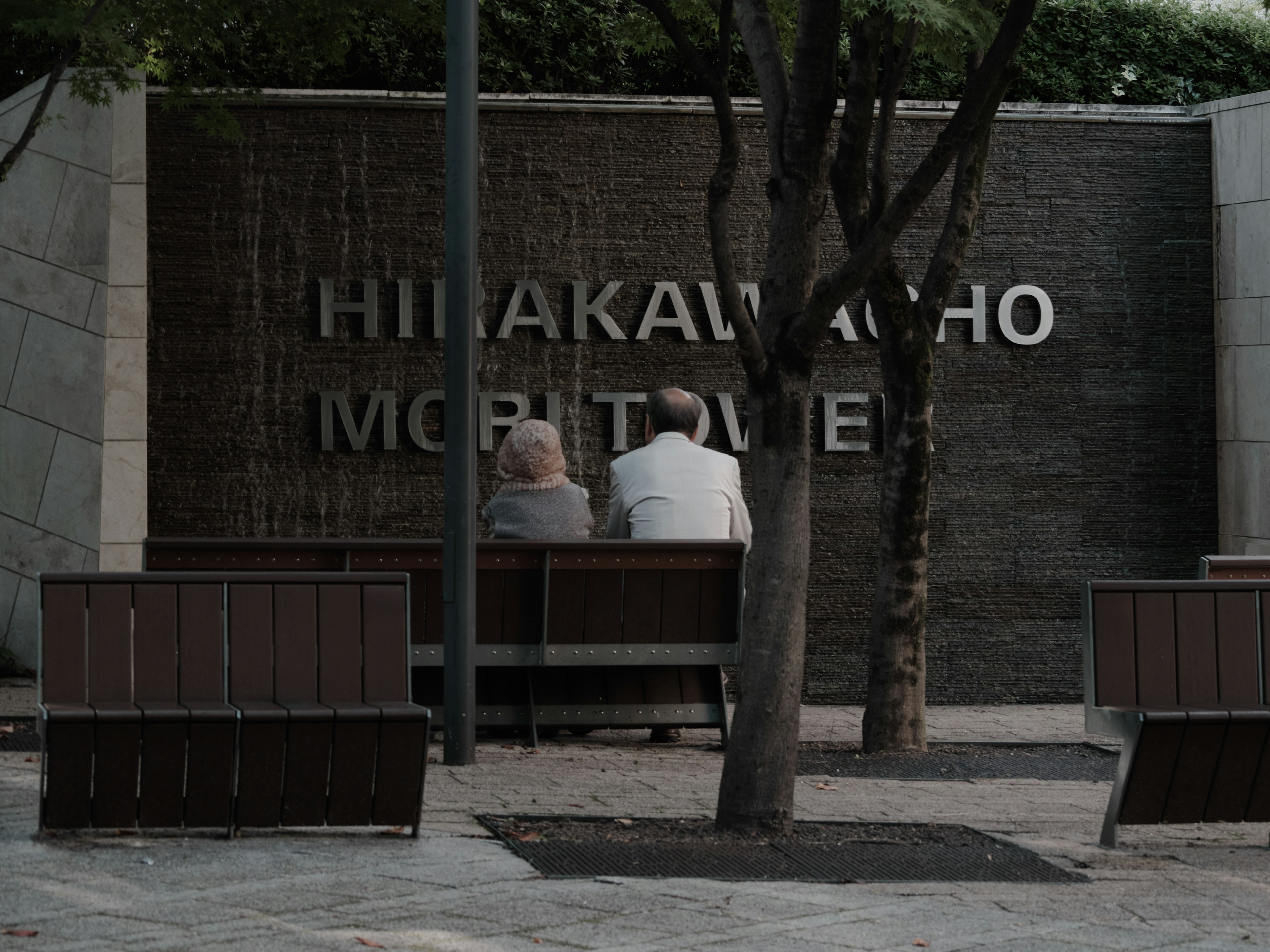 Zwei ältere Personen sitzen auf einer Bank vor dem Hinakawacho-Denkmal