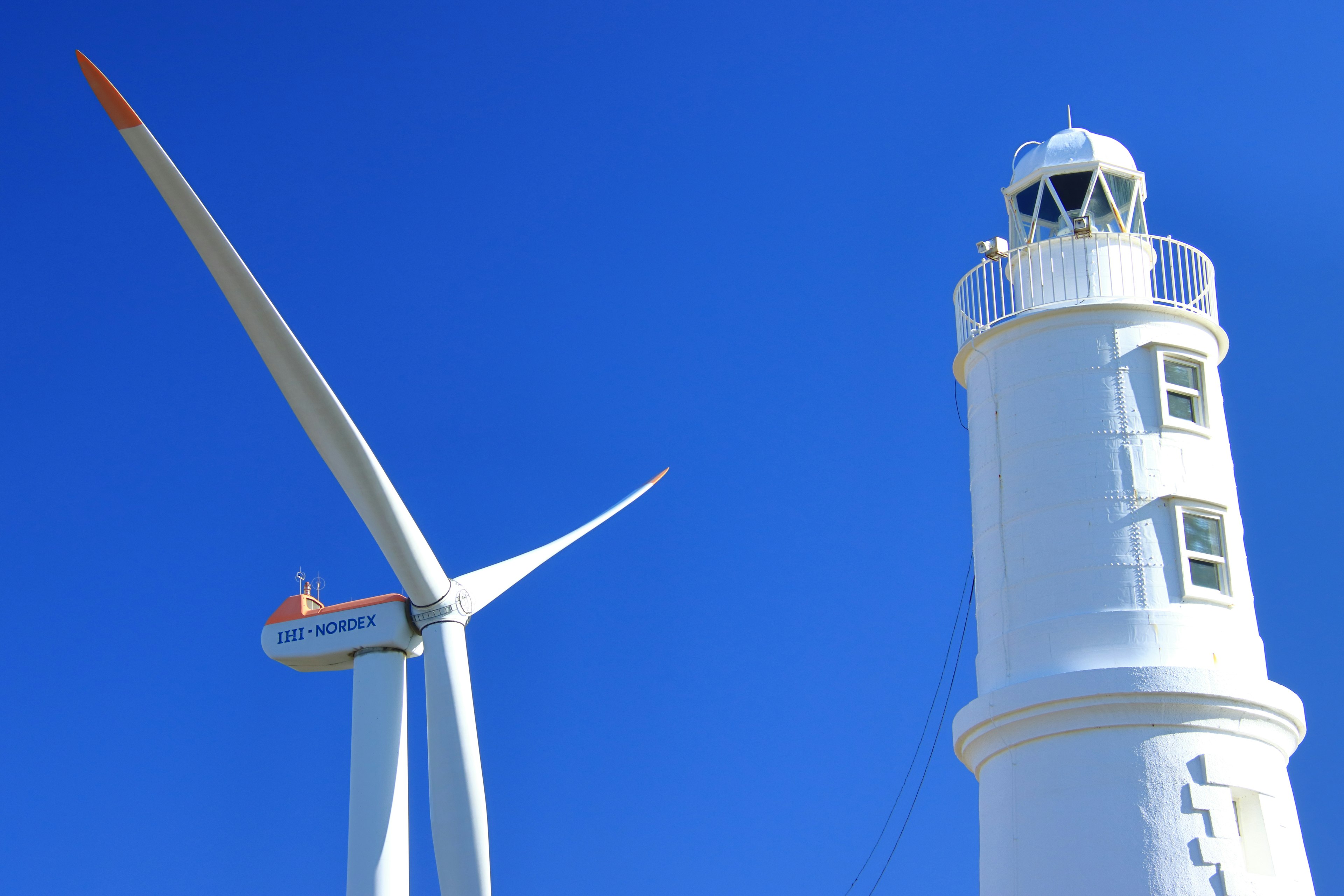 Wind turbine and white lighthouse standing under a blue sky