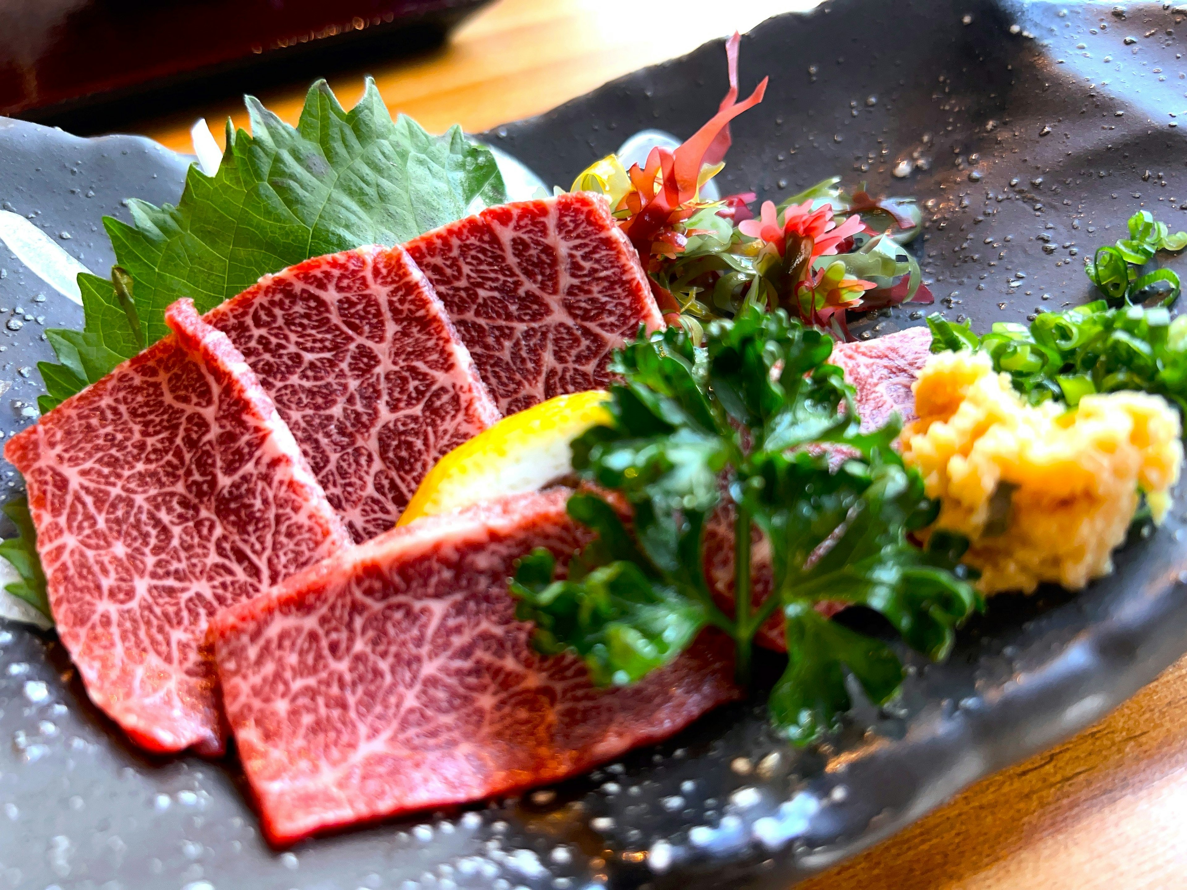 Sashimi de Wagyu joliment présenté avec des feuilles vertes et des condiments