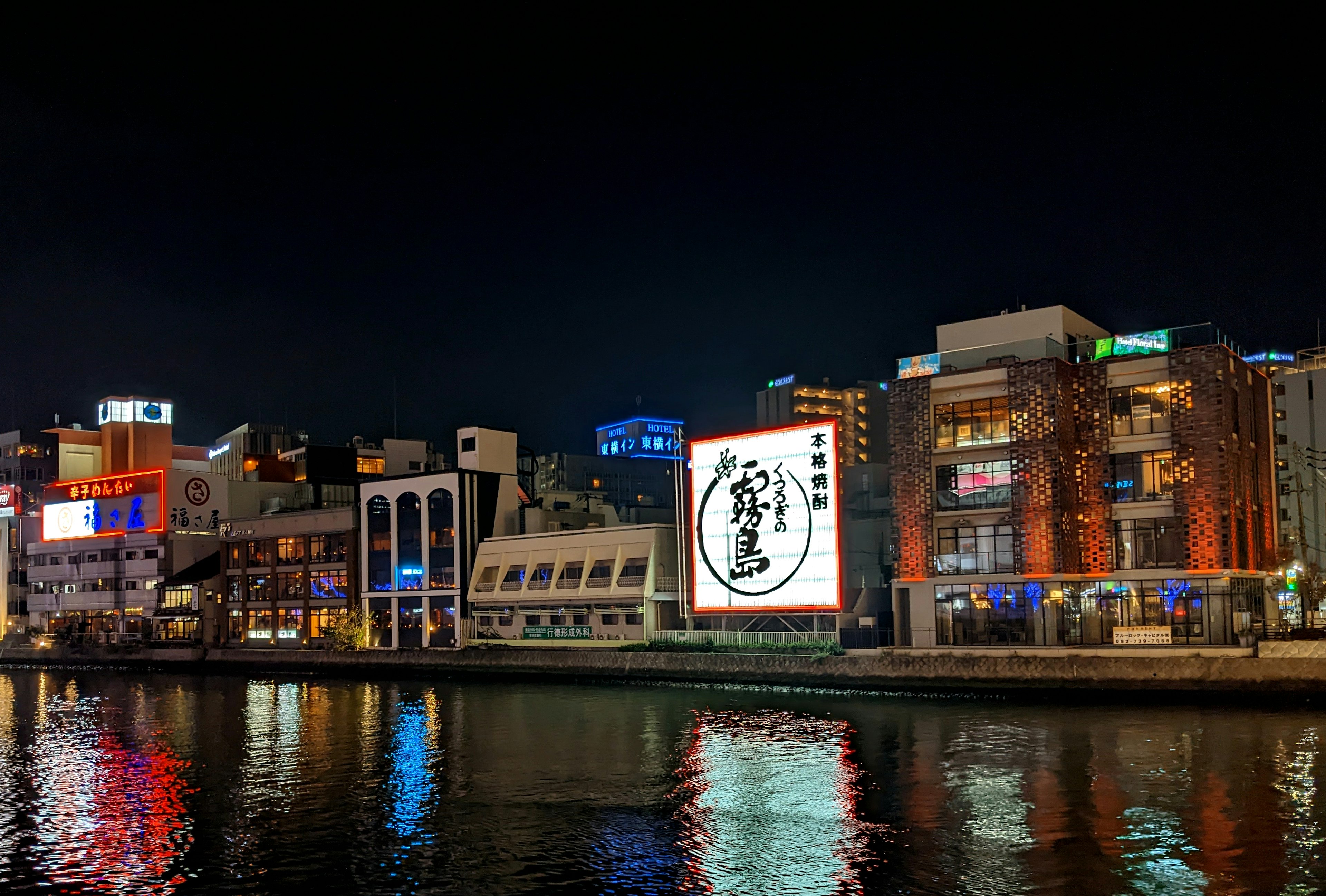 Escena urbana junto al agua de noche con edificios iluminados y letreros de neón