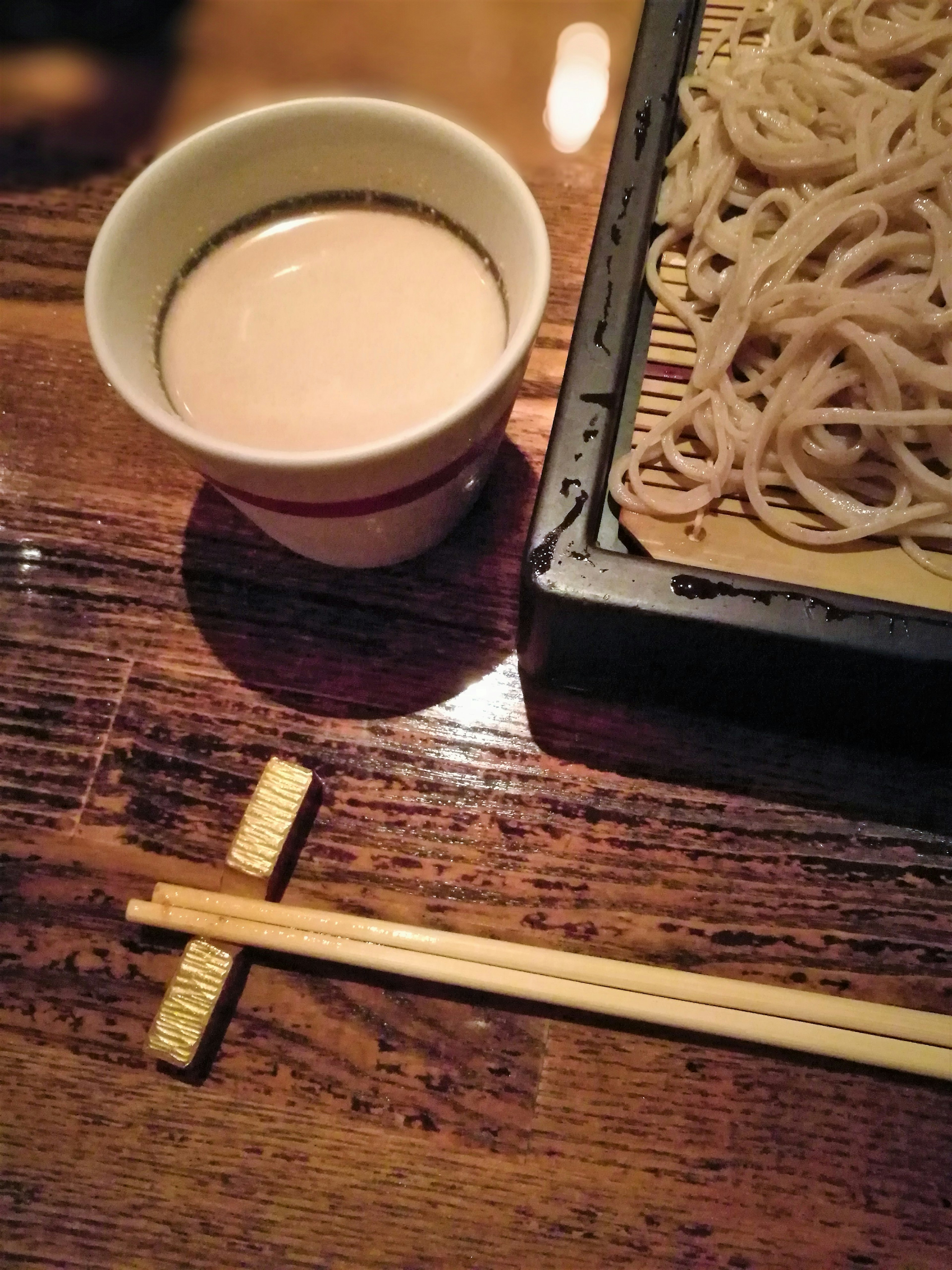 Sebuah cangkir keramik putih dengan minuman ringan di samping seporsi mie soba di atas meja kayu