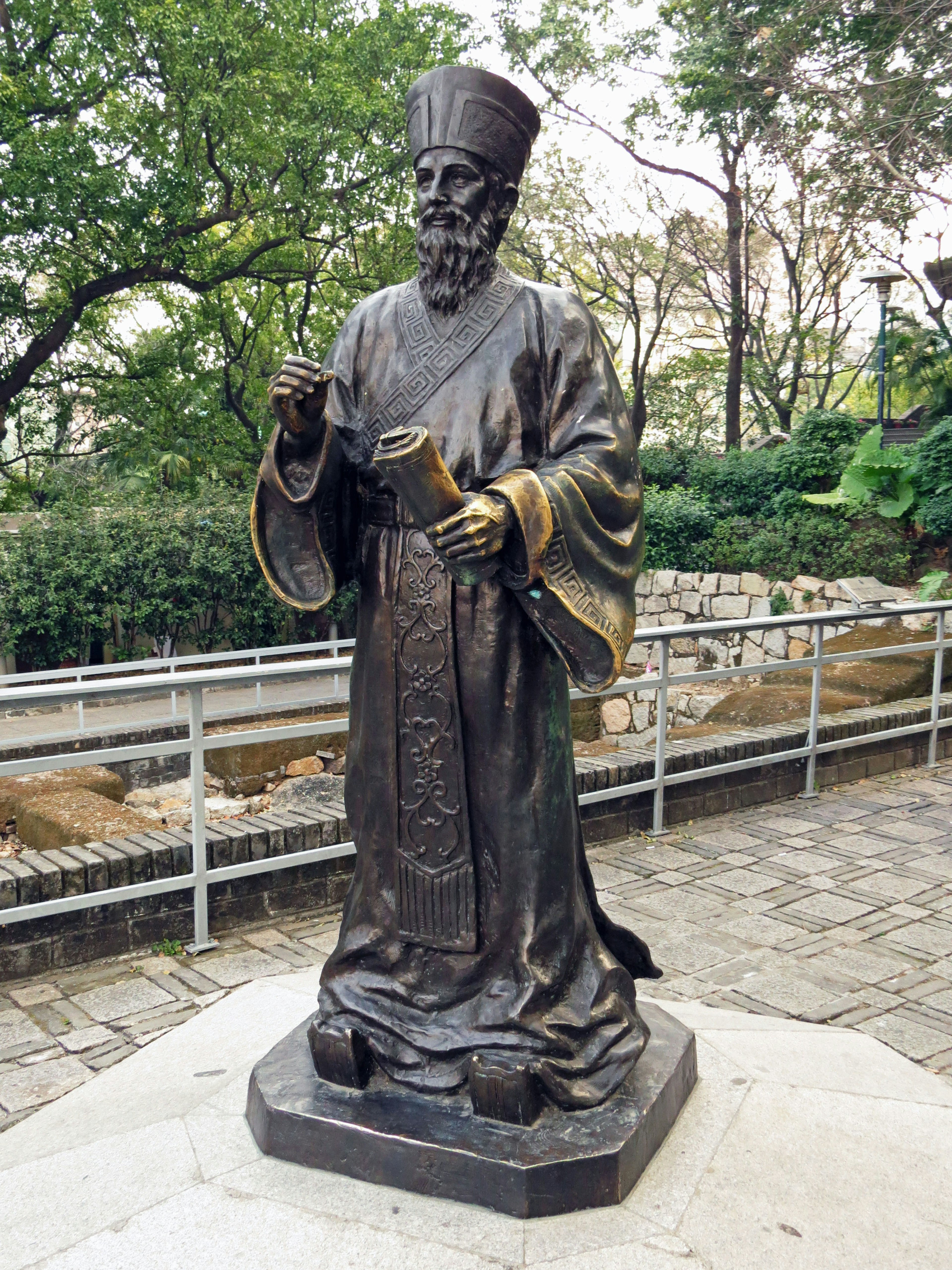 The statue depicts a figure in traditional attire holding a scroll with green trees in the background