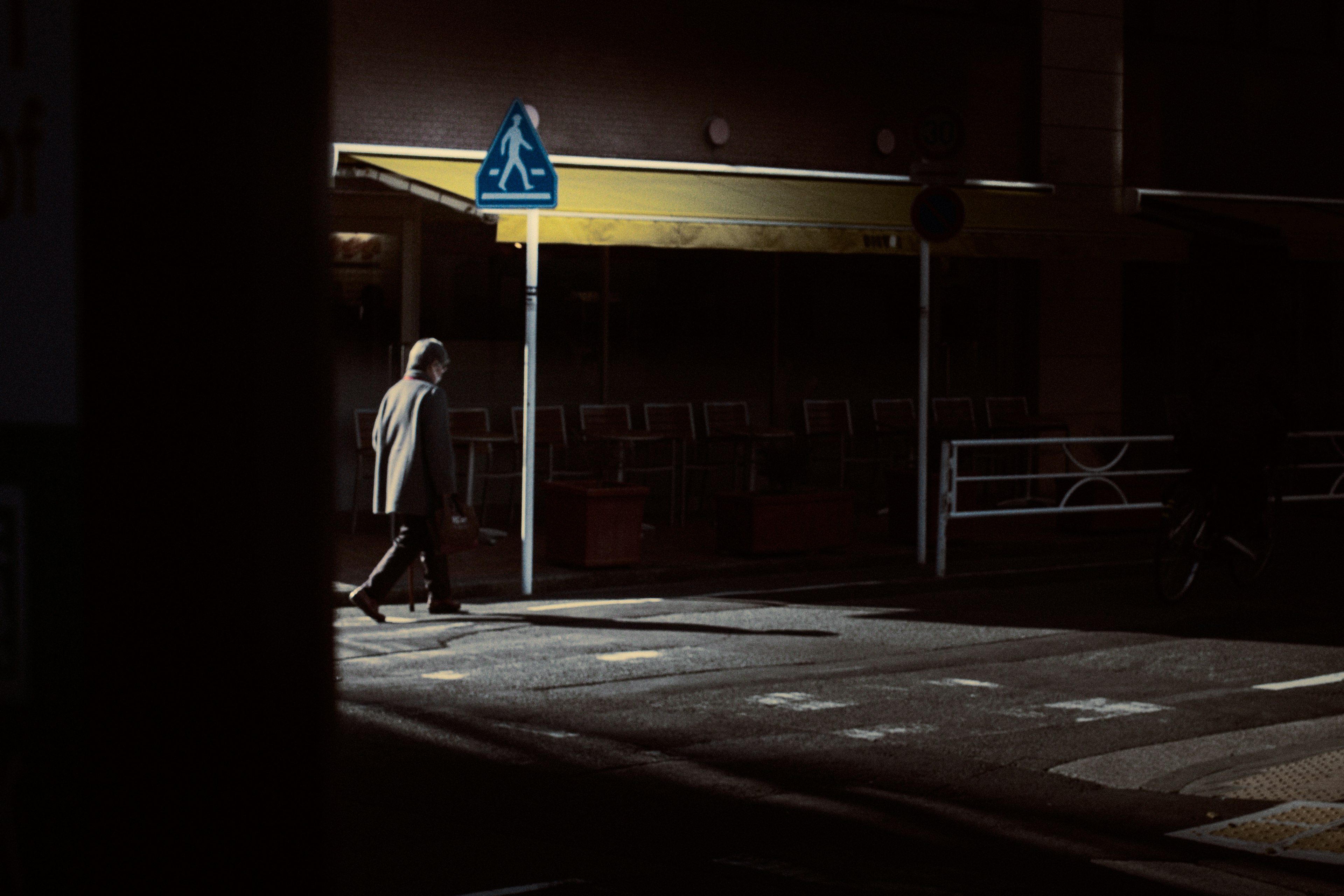 A person walking in a dimly lit street at night with a blue pedestrian sign