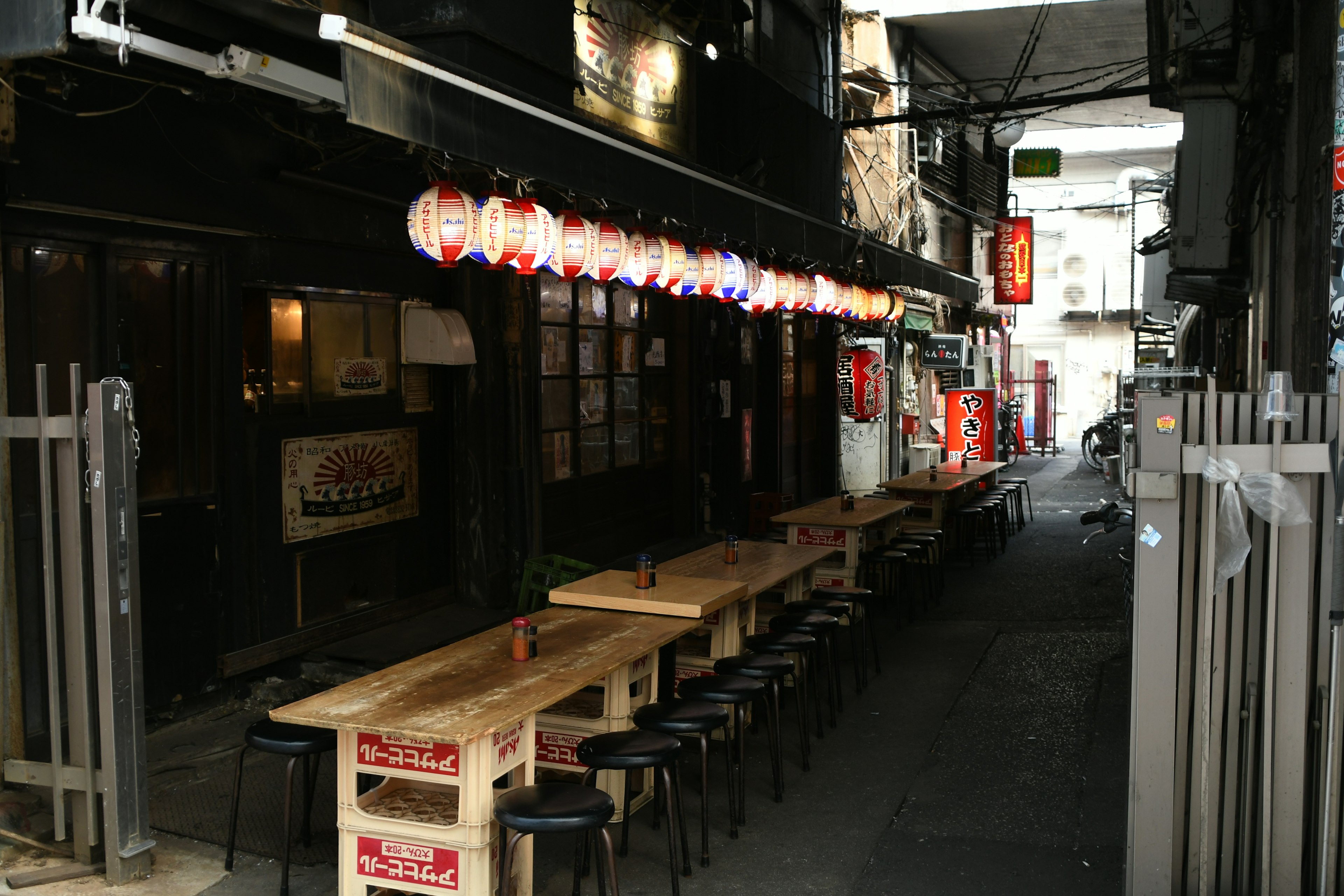 狭い路地に並ぶ木製テーブルと椅子 提灯が吊るされた飲食店の外観