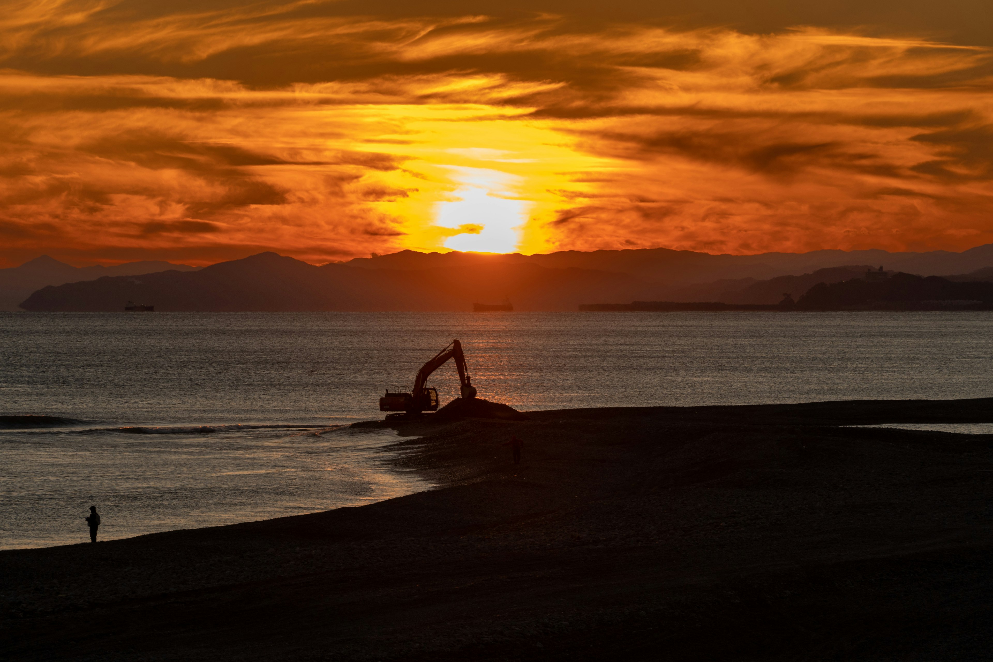 夕焼けの空の下で海に向かう重機と人影