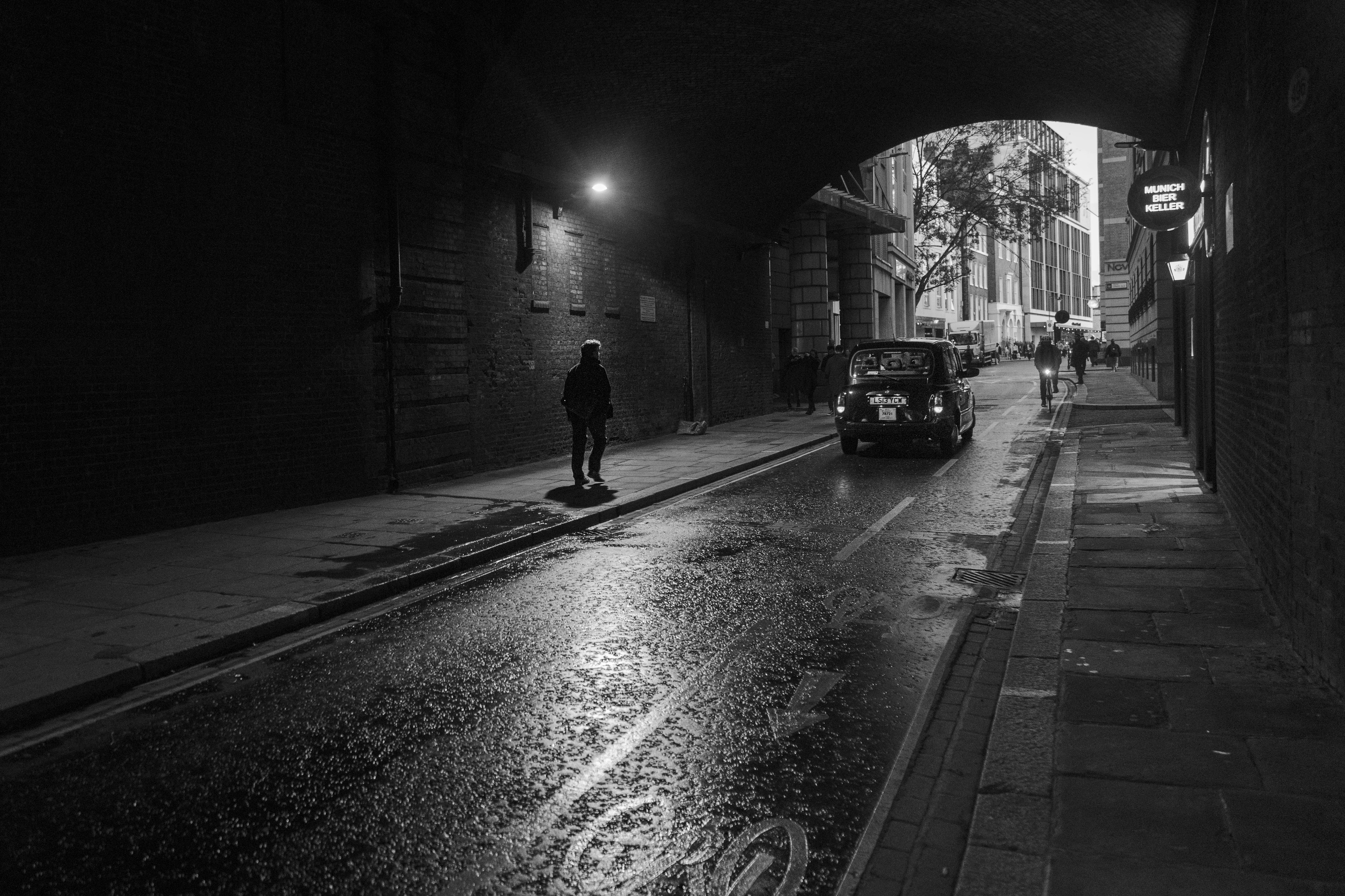 Una persona caminando bajo un arco oscuro con un coche pasando por una calle de la ciudad