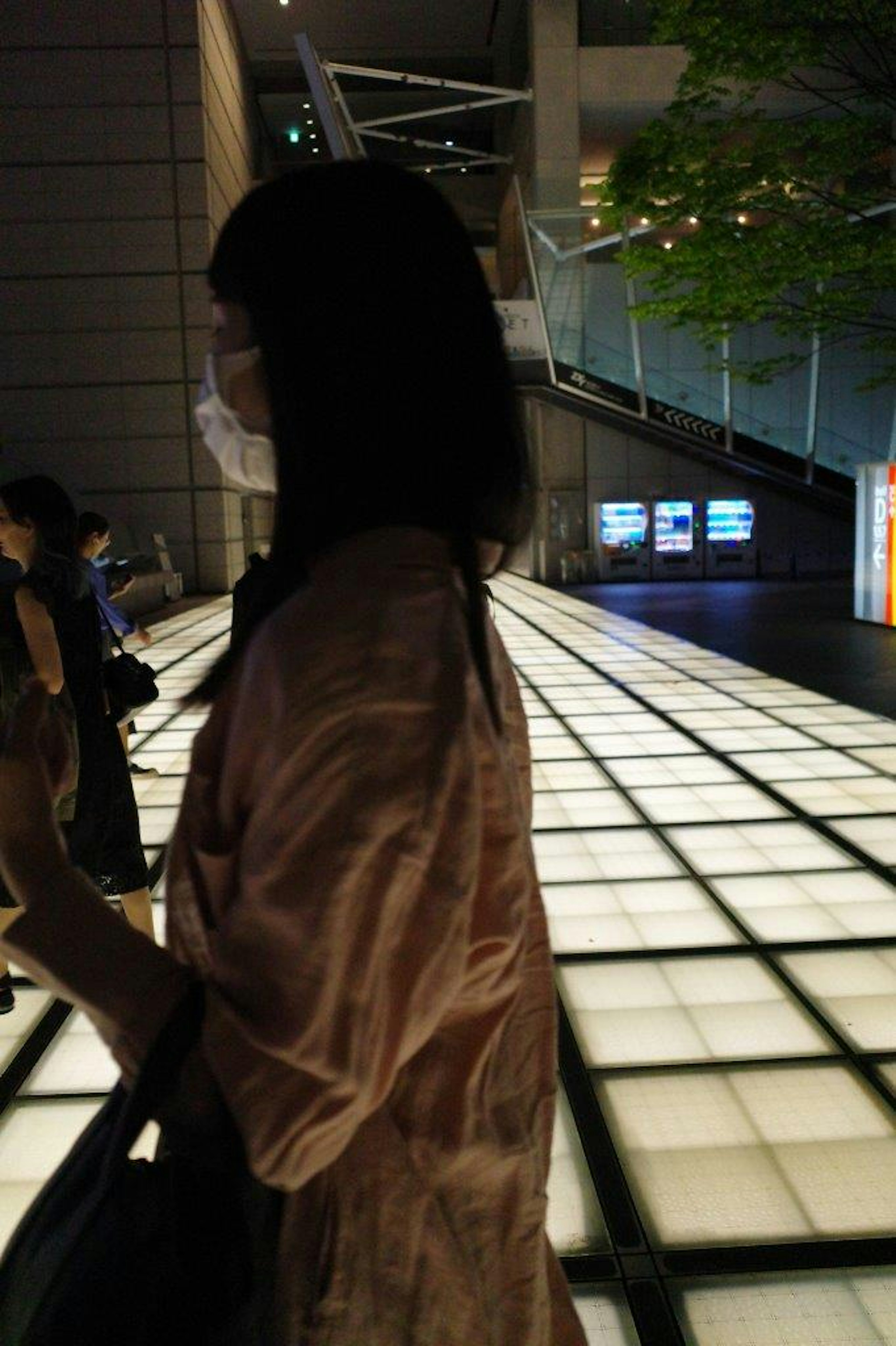 Una donna con una maschera sta in piedi di lato in un ambiente urbano notturno con un pavimento piastrellato luminoso