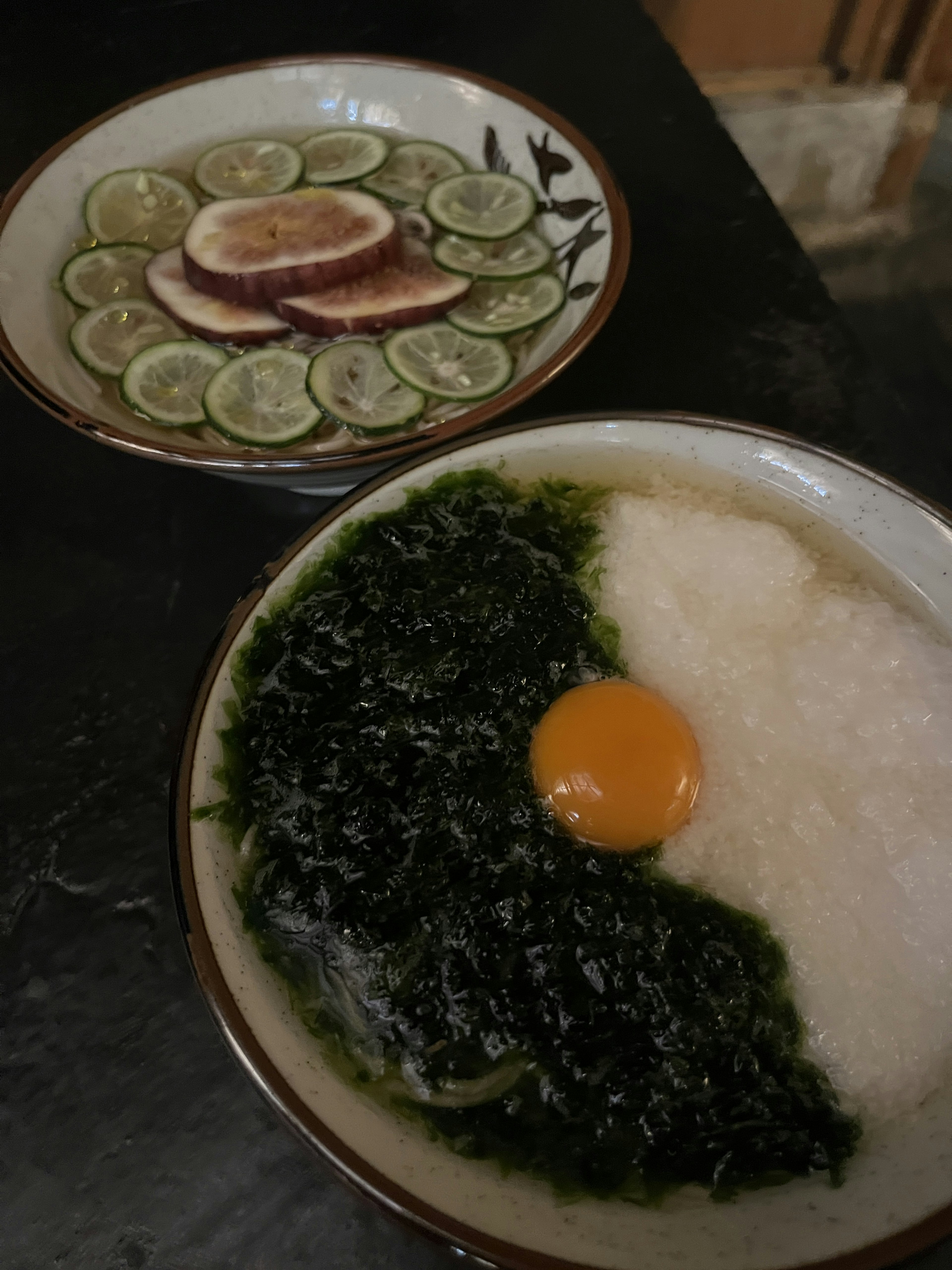 Semangkuk bubur nasi dengan rumput laut hijau dan telur di samping piring berisi buah sitrun yang diiris