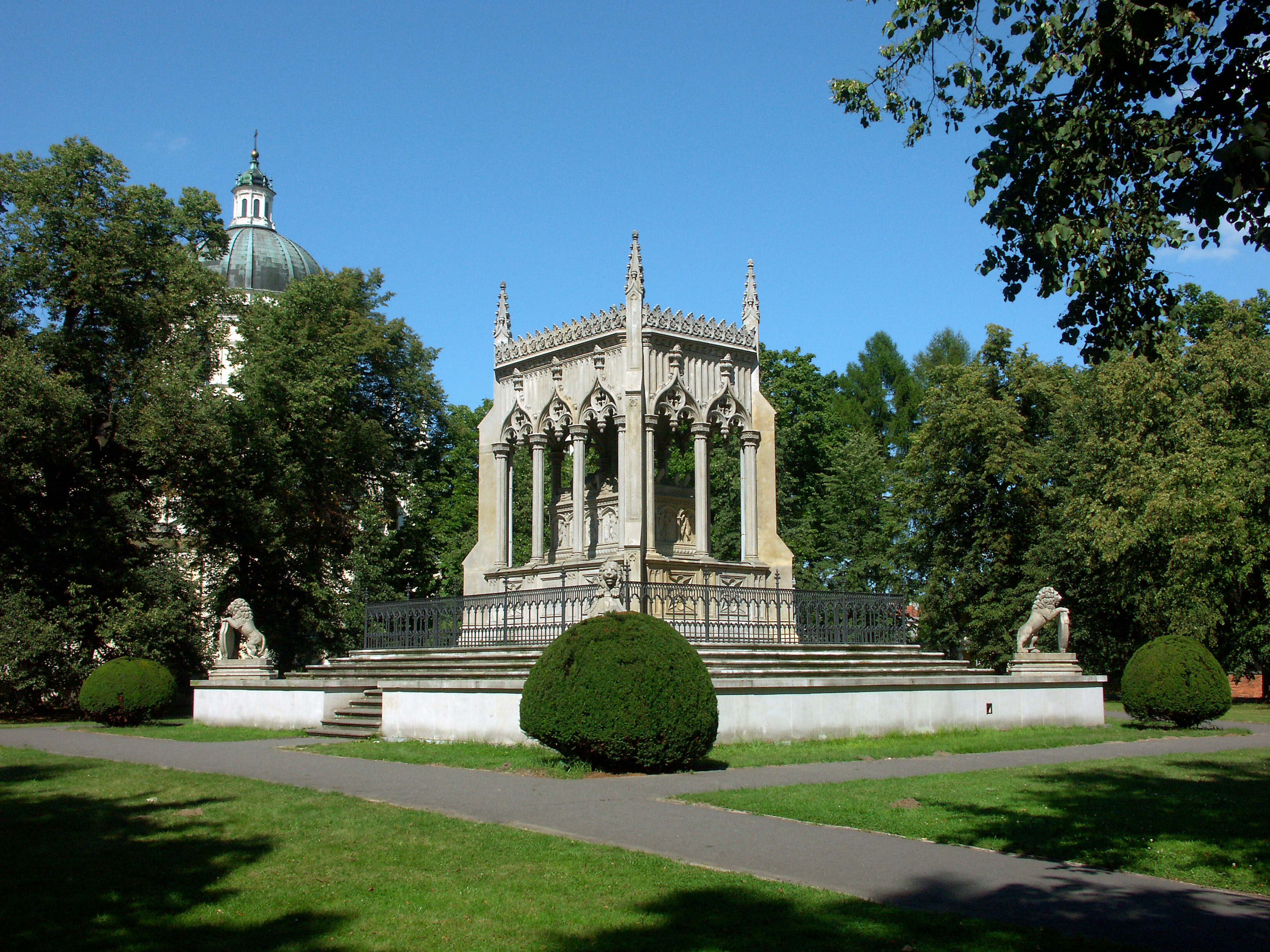 Steinpavillon in einem Park mit gestutzten Büschen unter einem klaren blauen Himmel