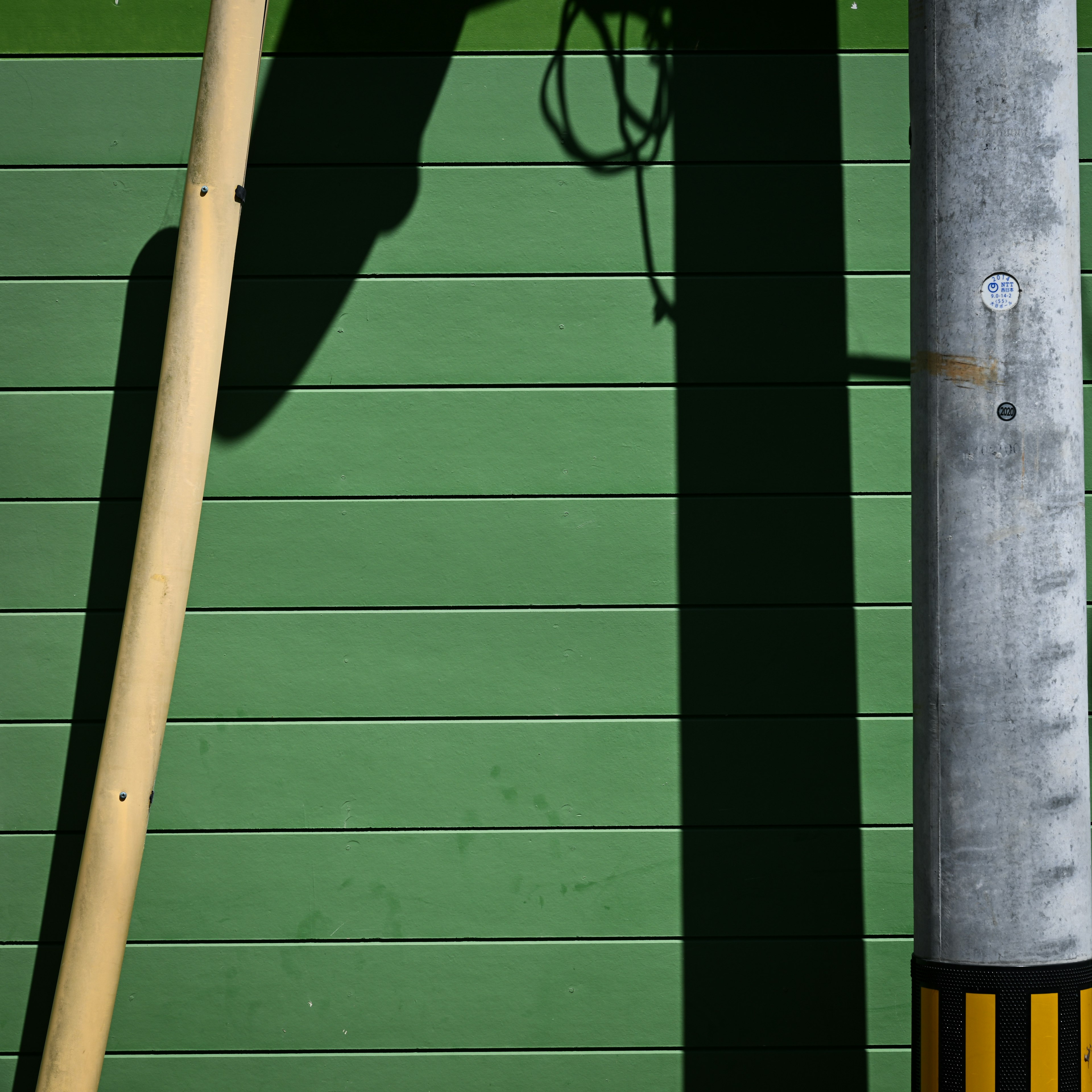 Schatten eines Holzpfostens und eines Metallpfostens an einer grünen Wand