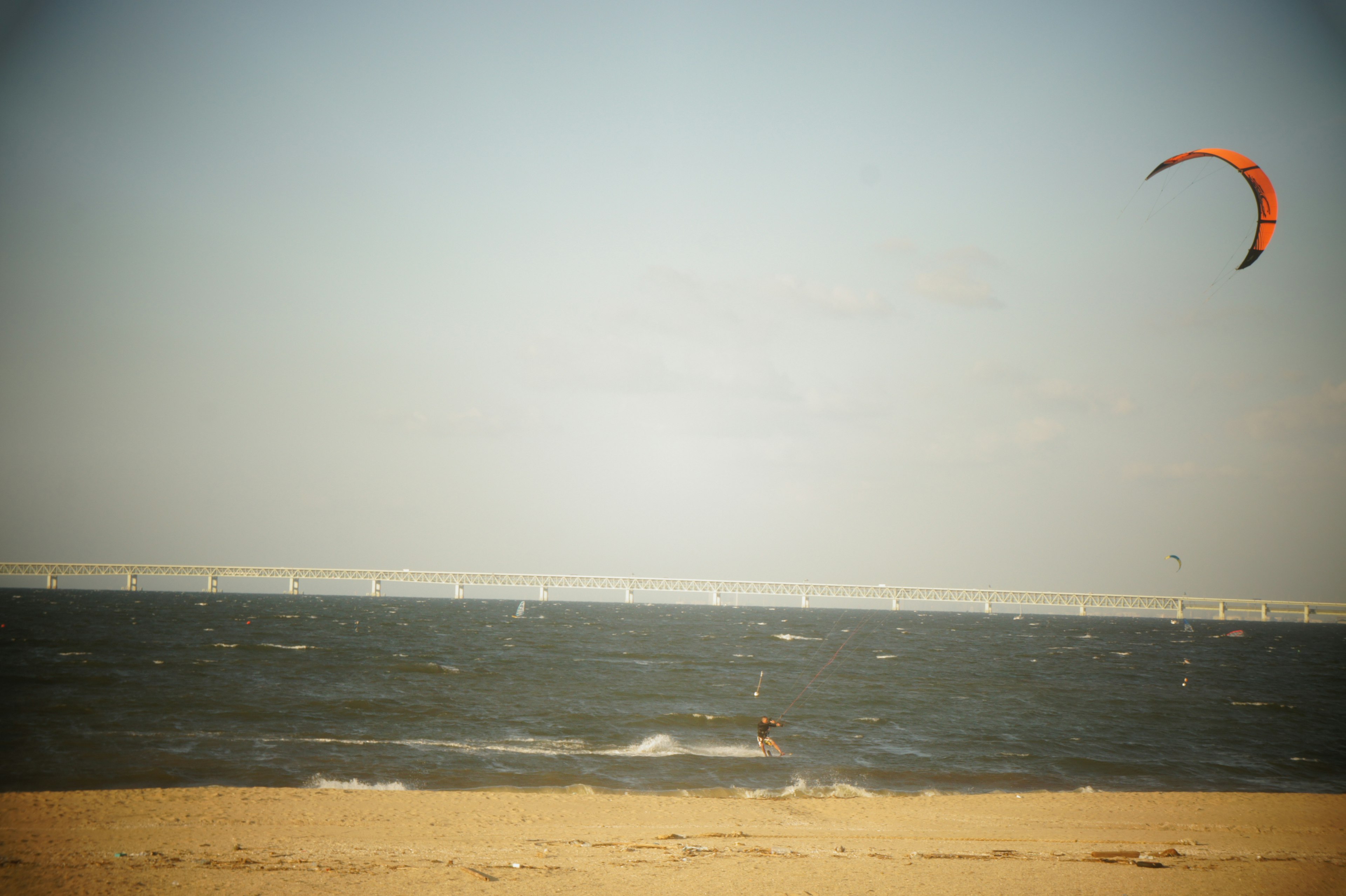 Eine Person beim Kitesurfen am Strand mit einer Brücke in der Ferne