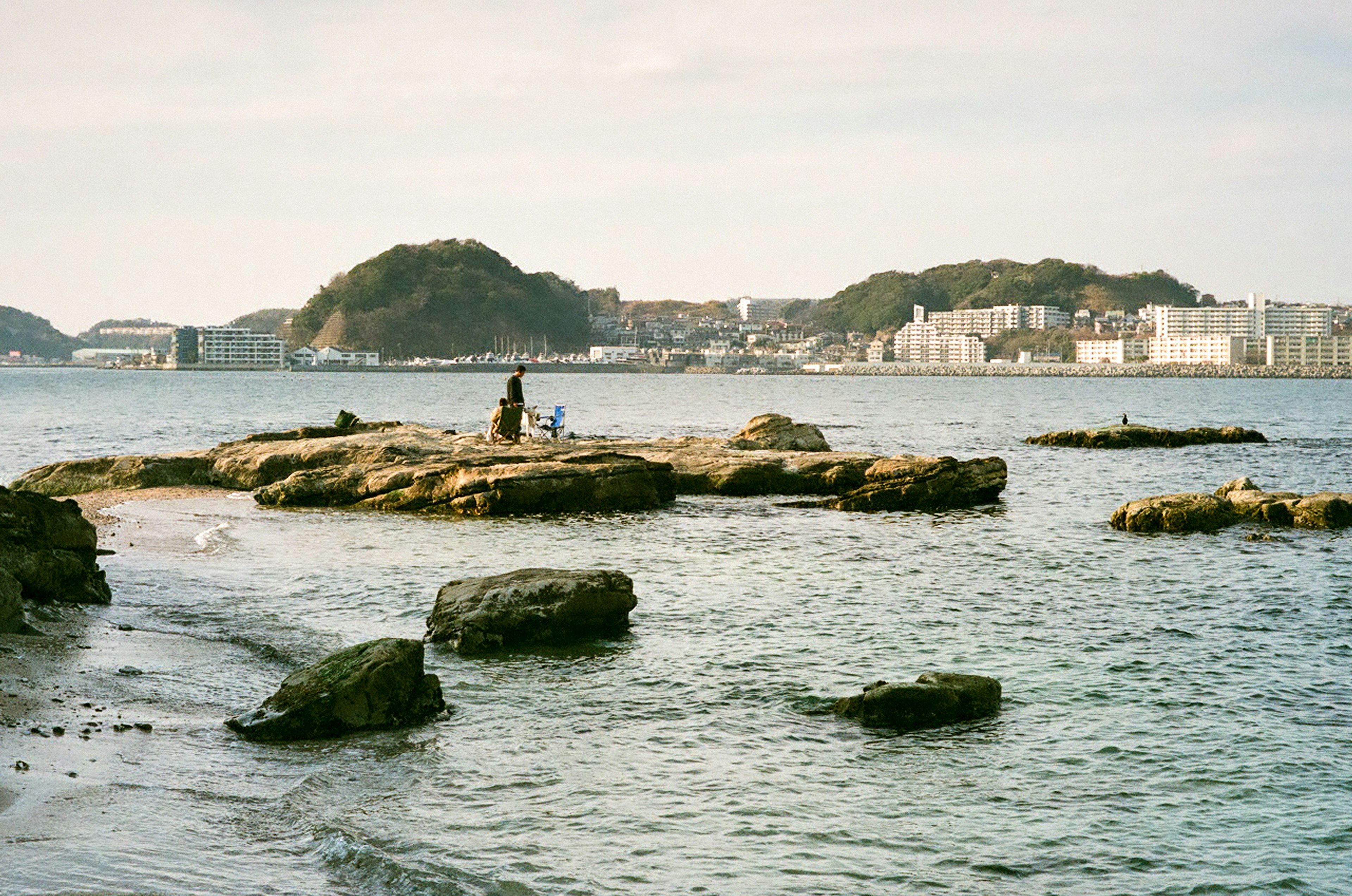 People fishing on rocky shore with calm water and distant hills