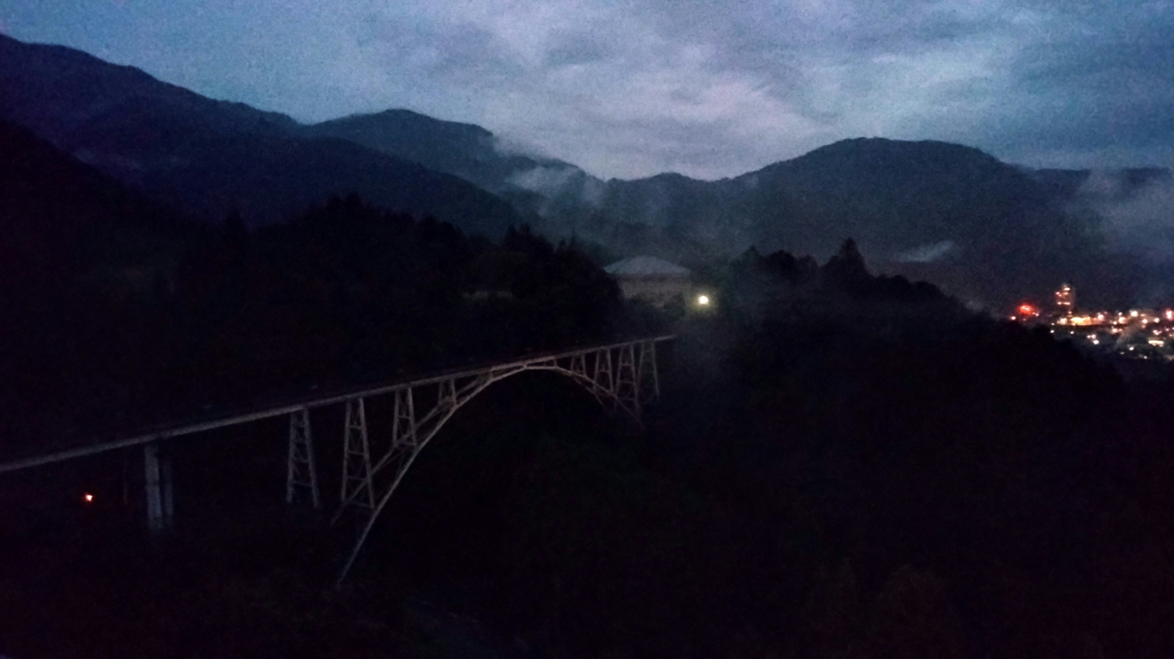 Jembatan busur dikelilingi pegunungan di malam hari dengan lampu kota di kejauhan