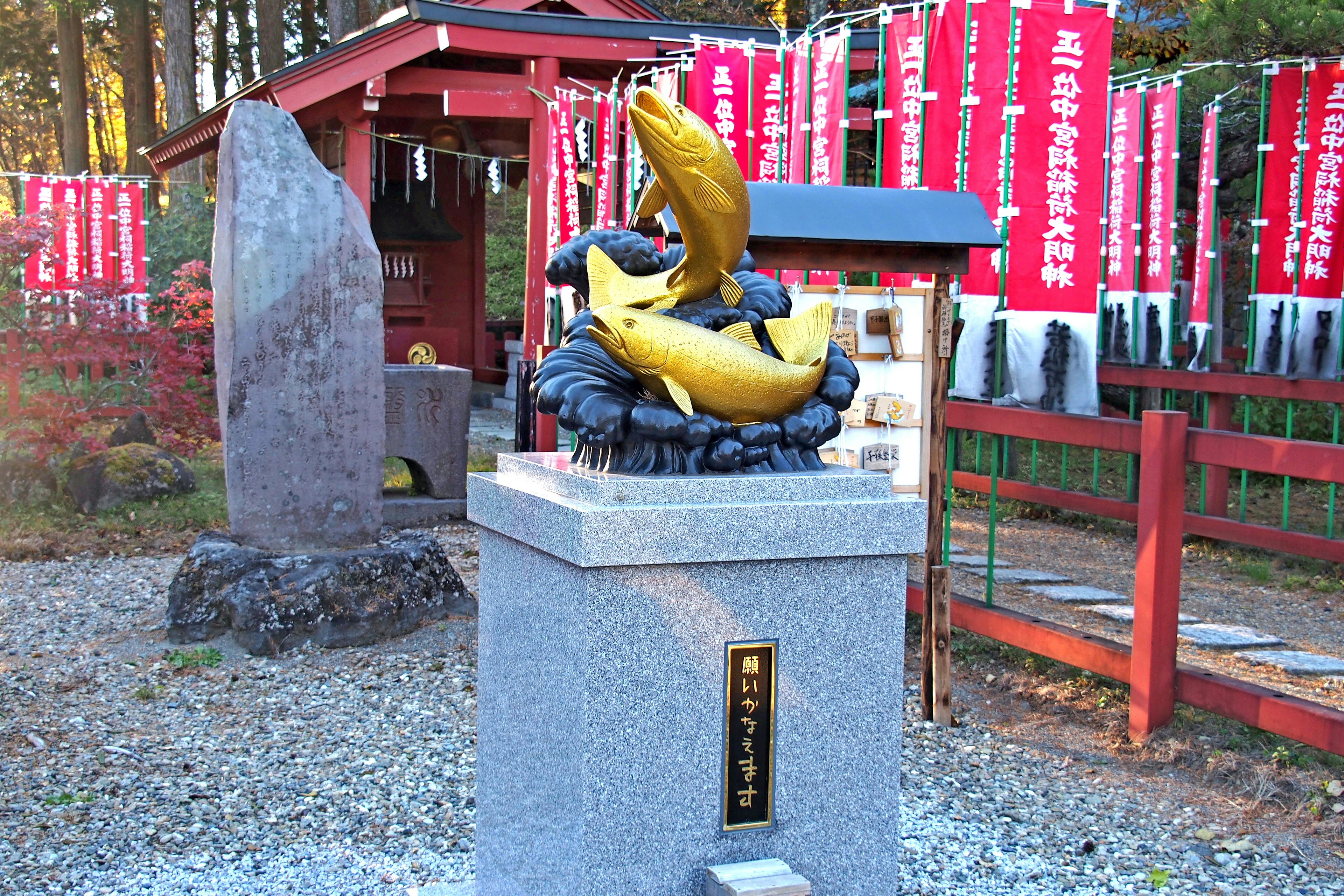 Eine goldene Fischskulptur in einer Schreinanlage mit roten Bannern im Hintergrund auf einem Steinsockel