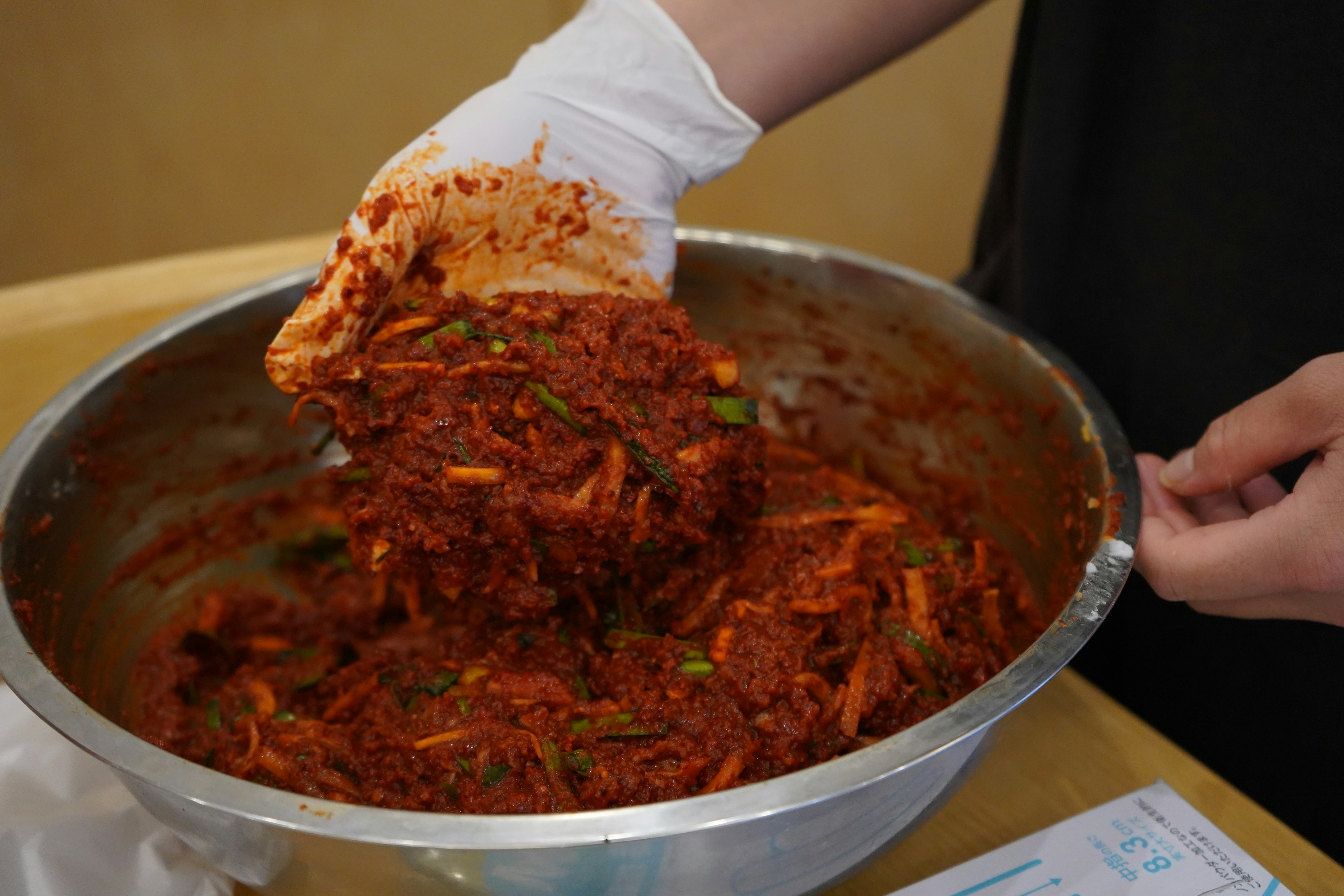 Una mano con guante mezclando ingredientes picantes de kimchi en un tazón grande