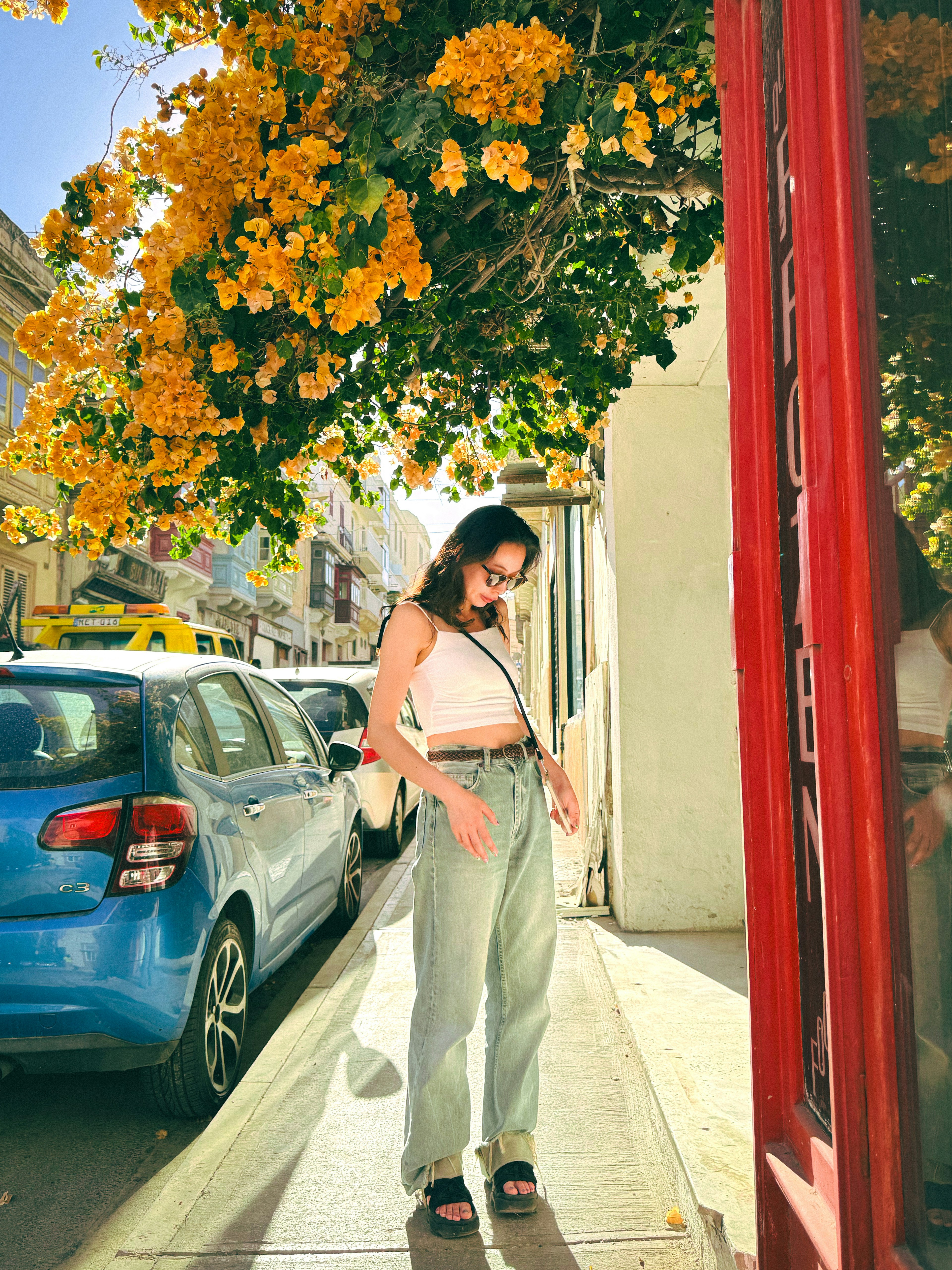 Woman standing under a yellow flowering tree next to a blue car