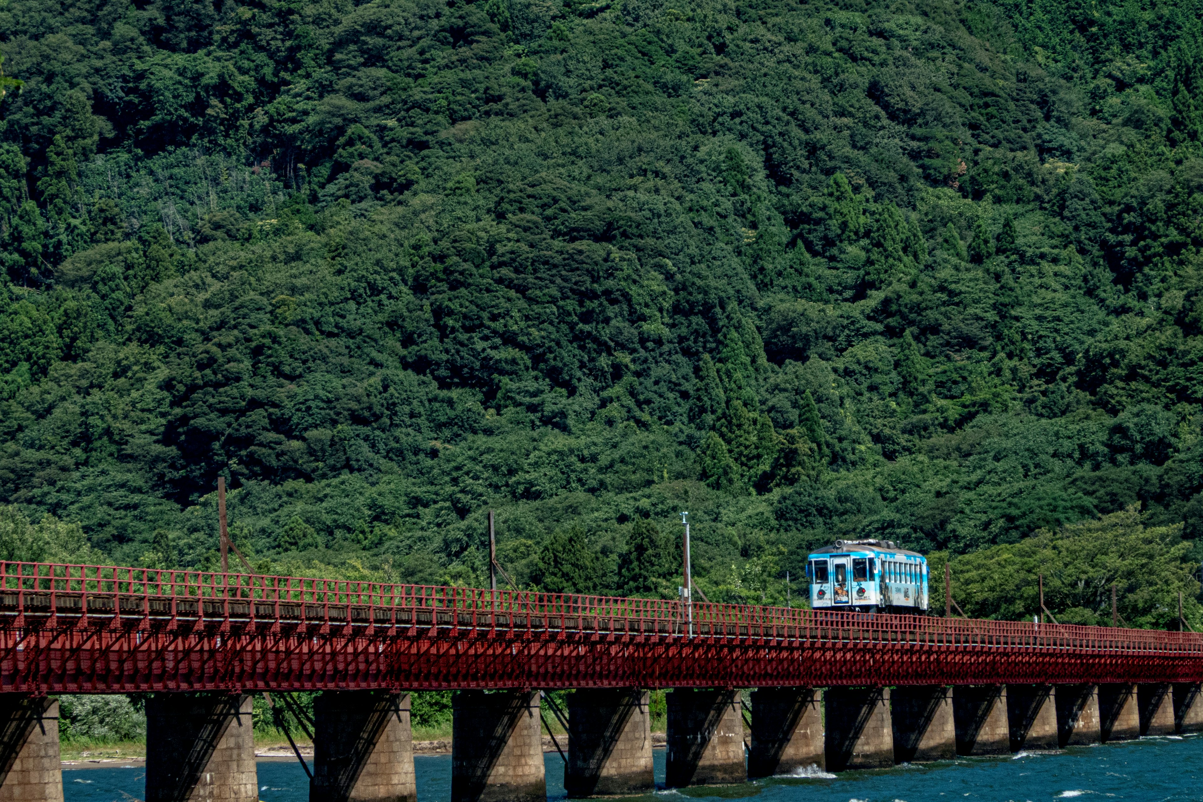 Blue train crossing a red bridge with green mountains in the background