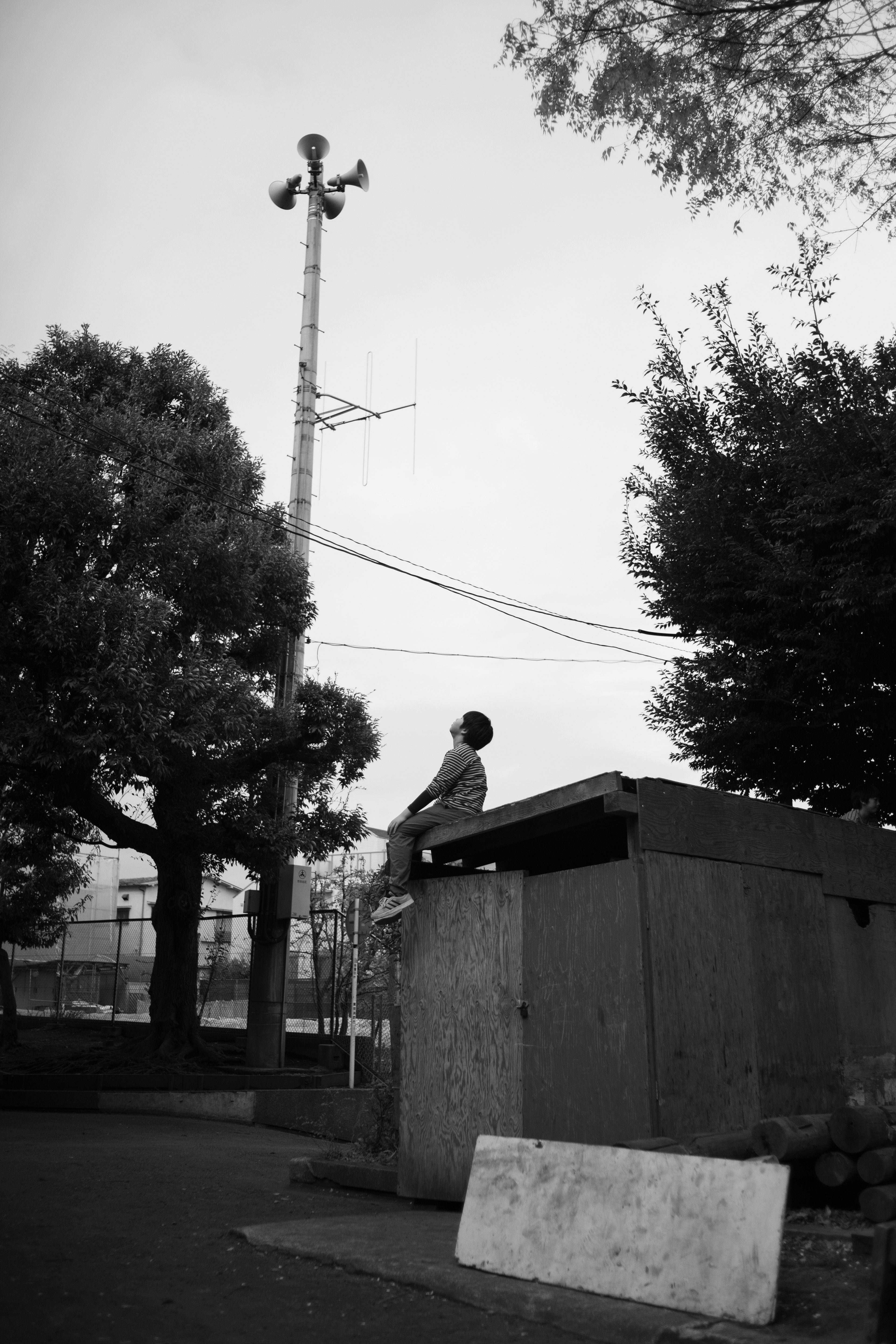 A black and white scene with a man sitting on top of a shed. In the background, a tall communication tower and trees are visible