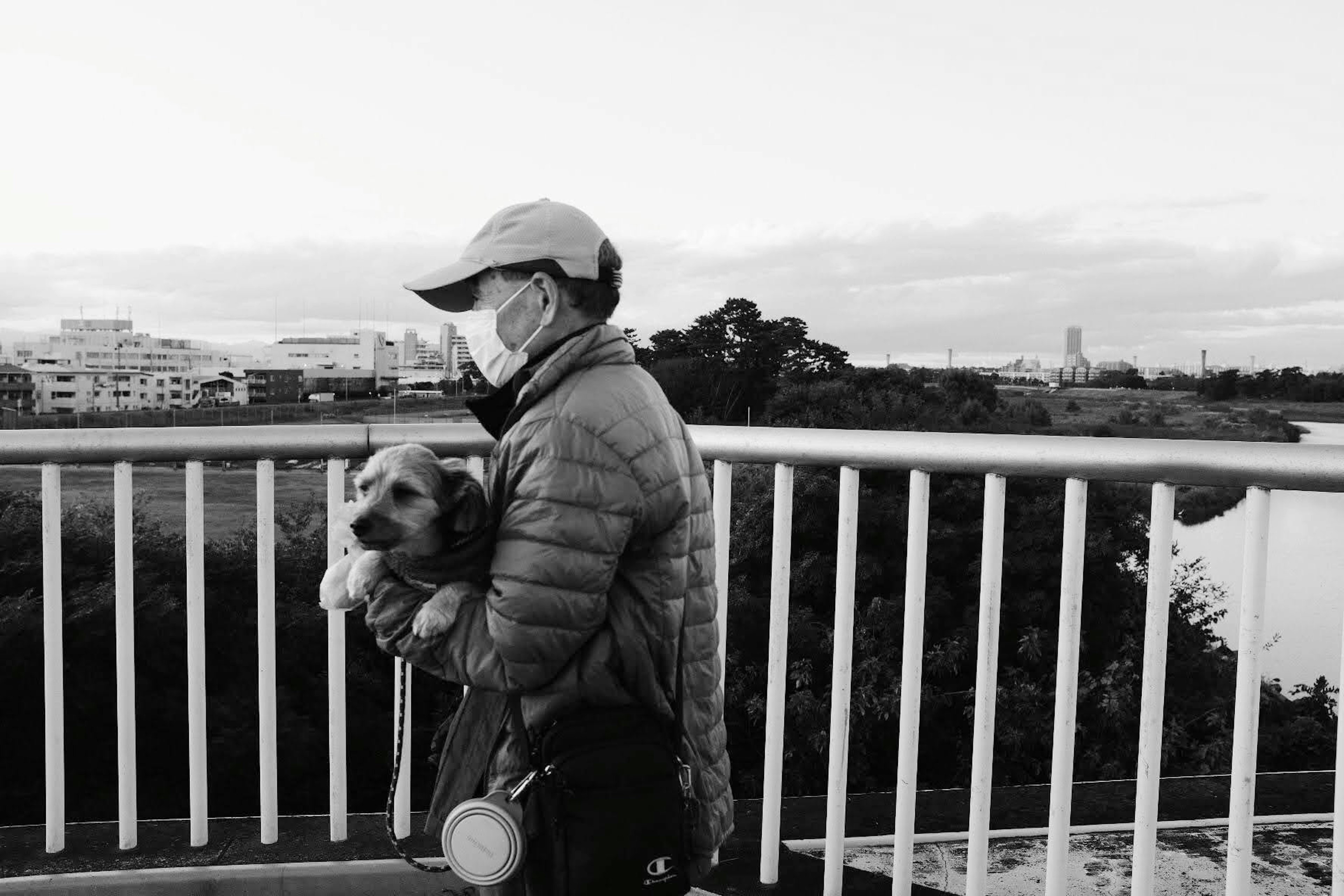 白黒の風景の中で犬を抱えた人が橋の上に立っている