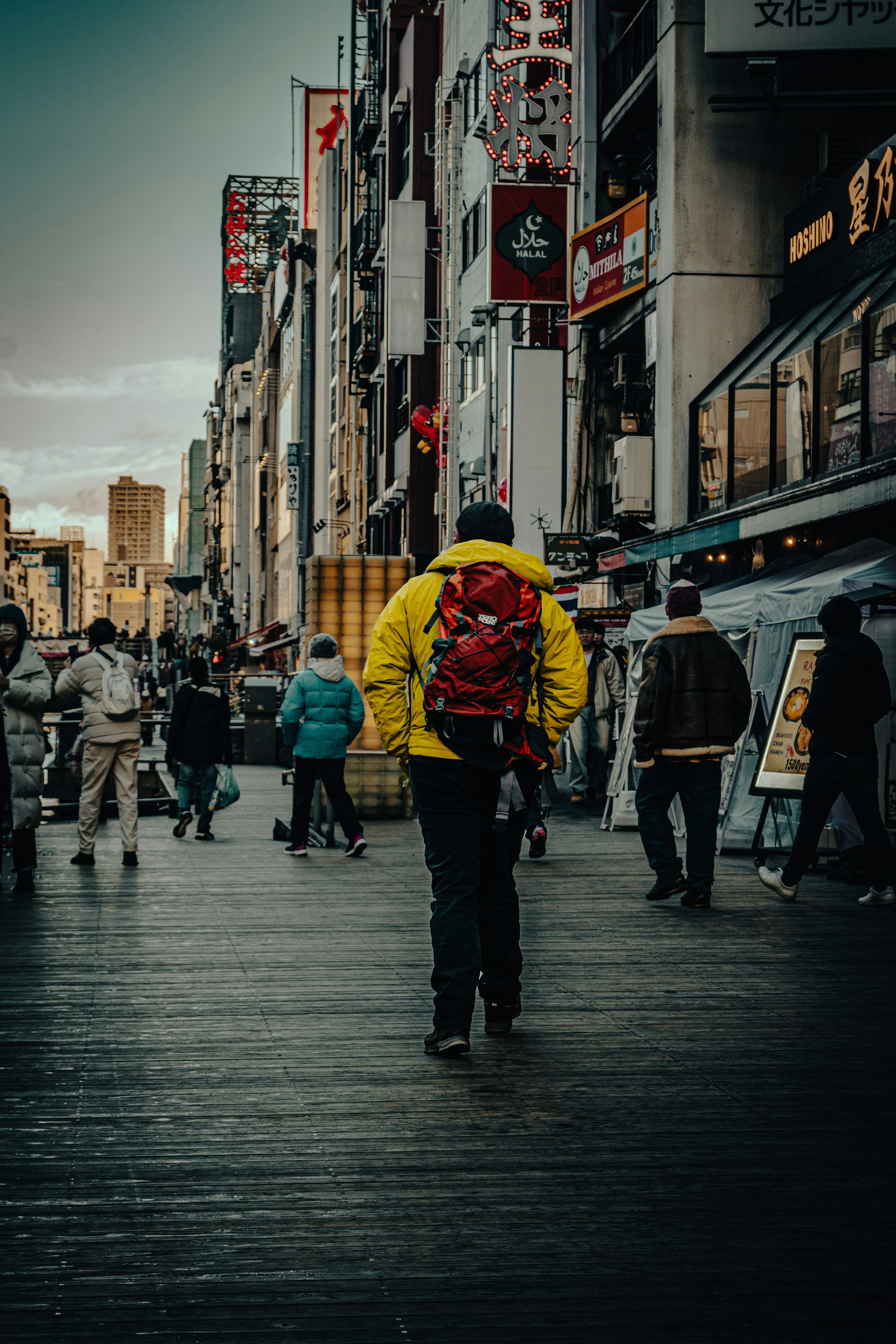 A man in a yellow jacket with a red backpack walking through a bustling street filled with people