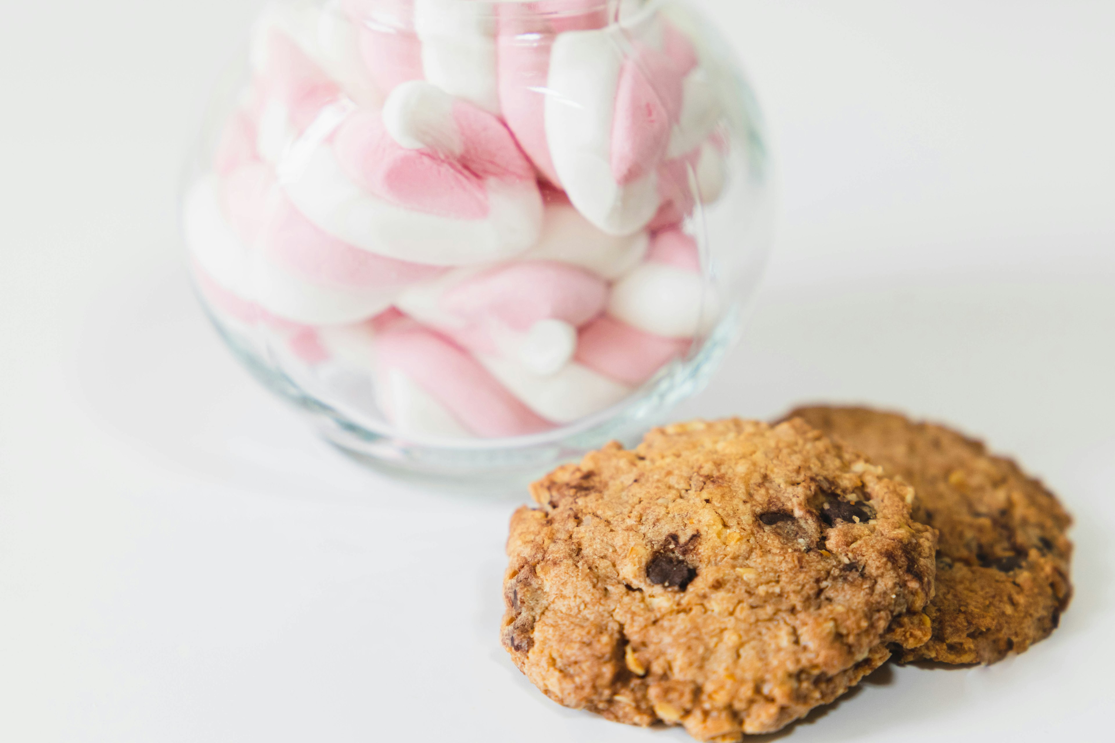 A glass jar filled with pink and white marshmallows alongside two chocolate chip cookies