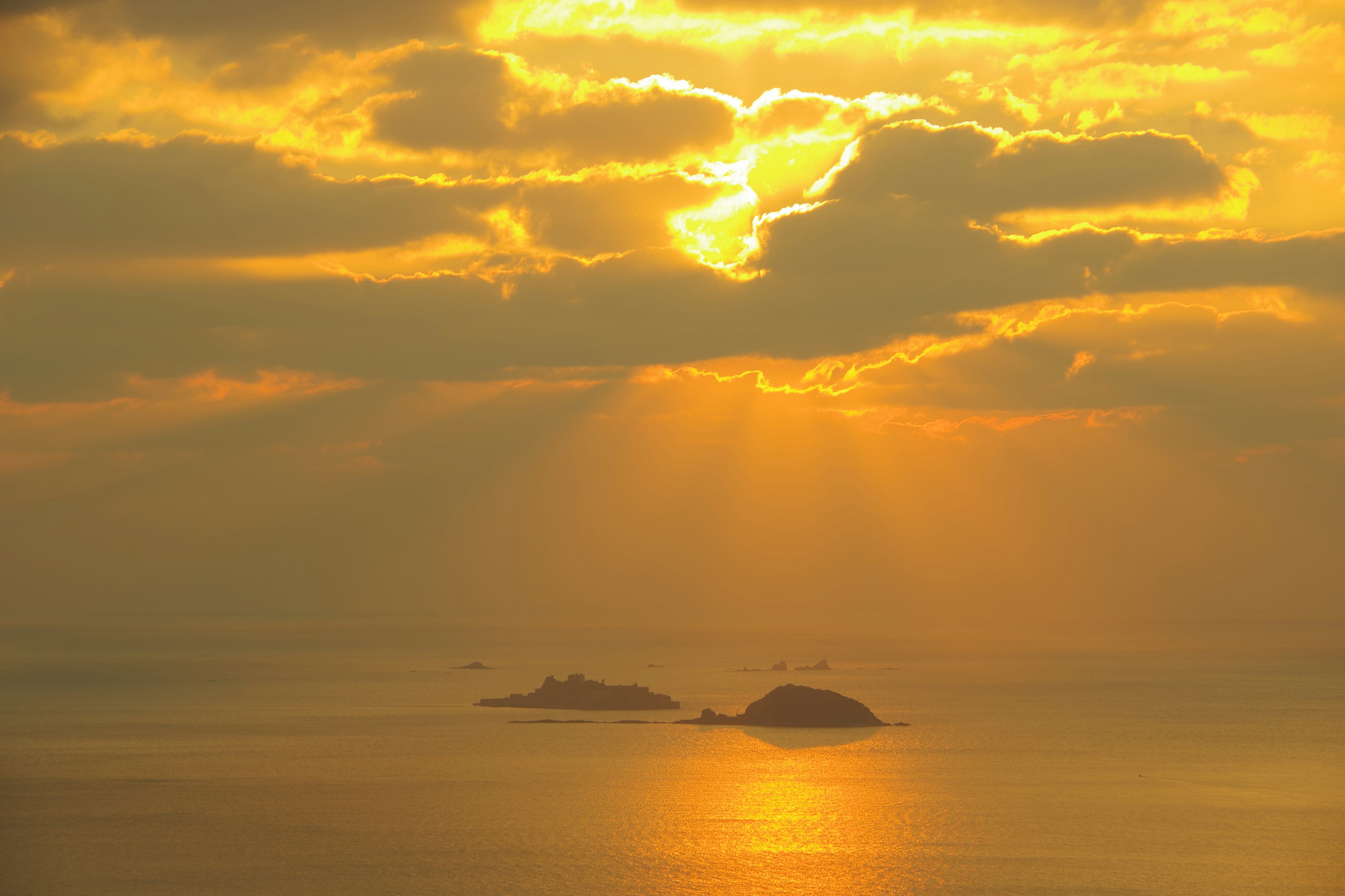 Raggi di sole dorati al tramonto che attraversano le nuvole riflettendosi su un mare calmo con silhouette di isole