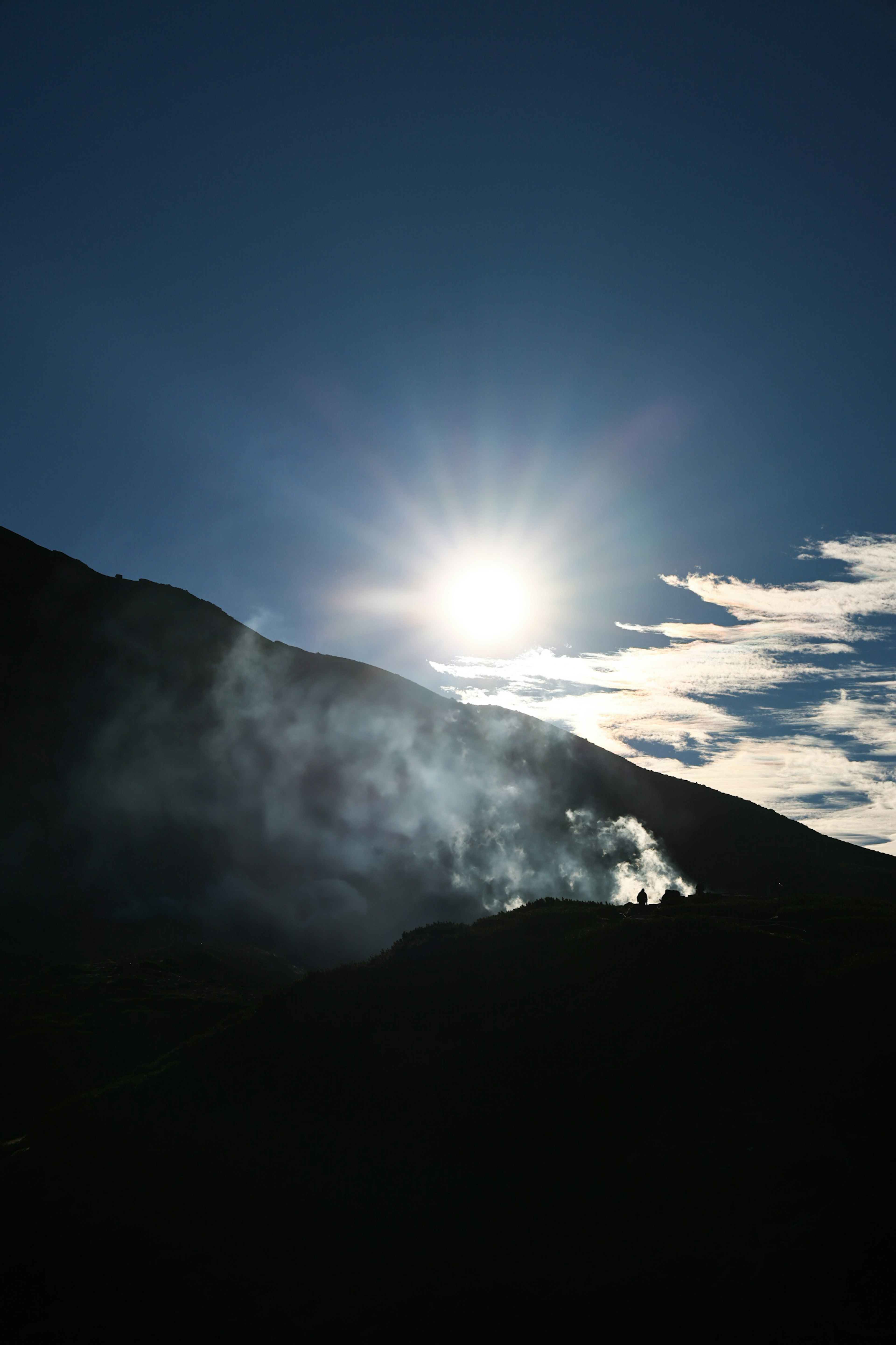 Silhouette gunung yang mengeluarkan asap melawan langit cerah dengan matahari