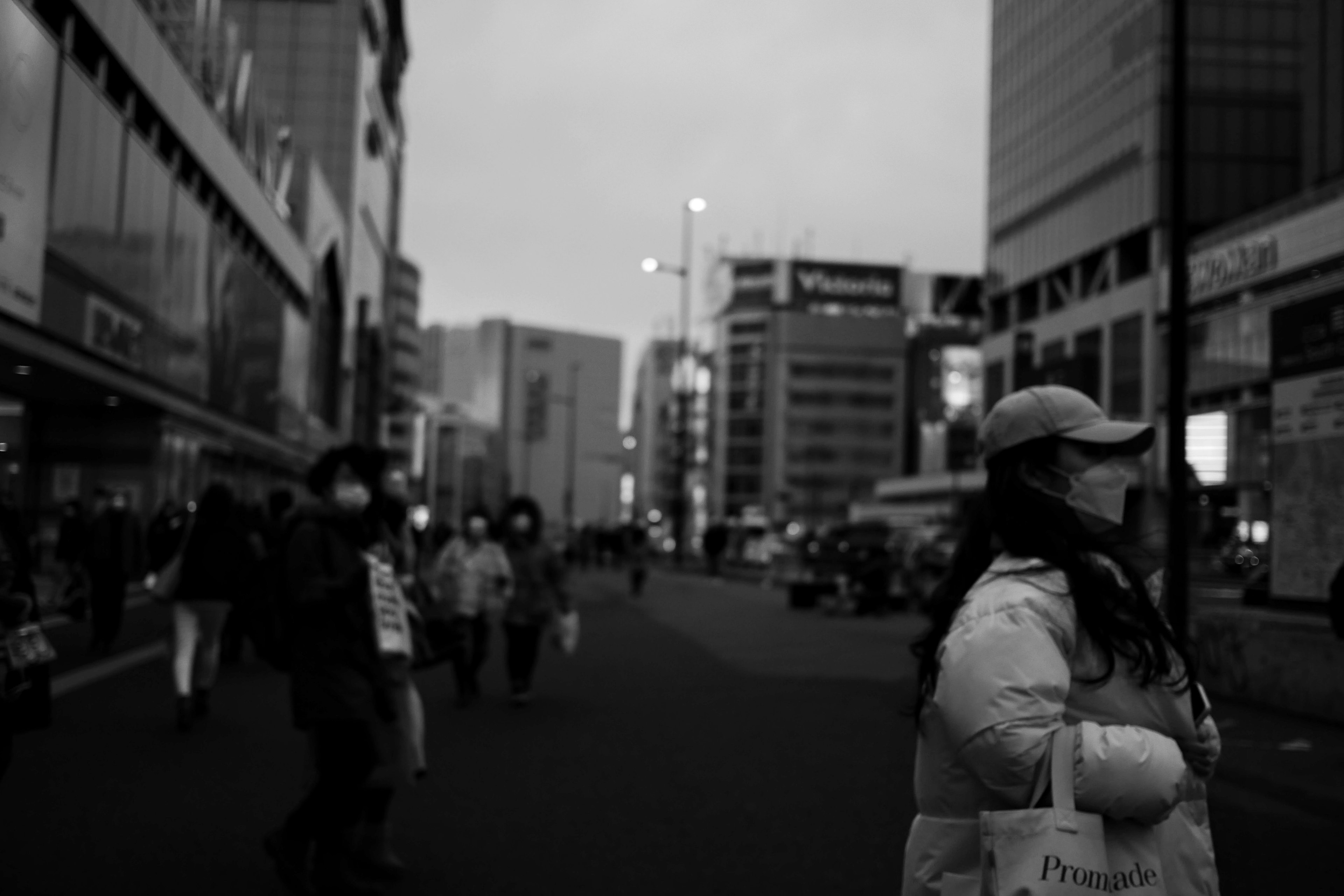 一名戴口罩的女性站在黑白城市街道上，周围有行人走过