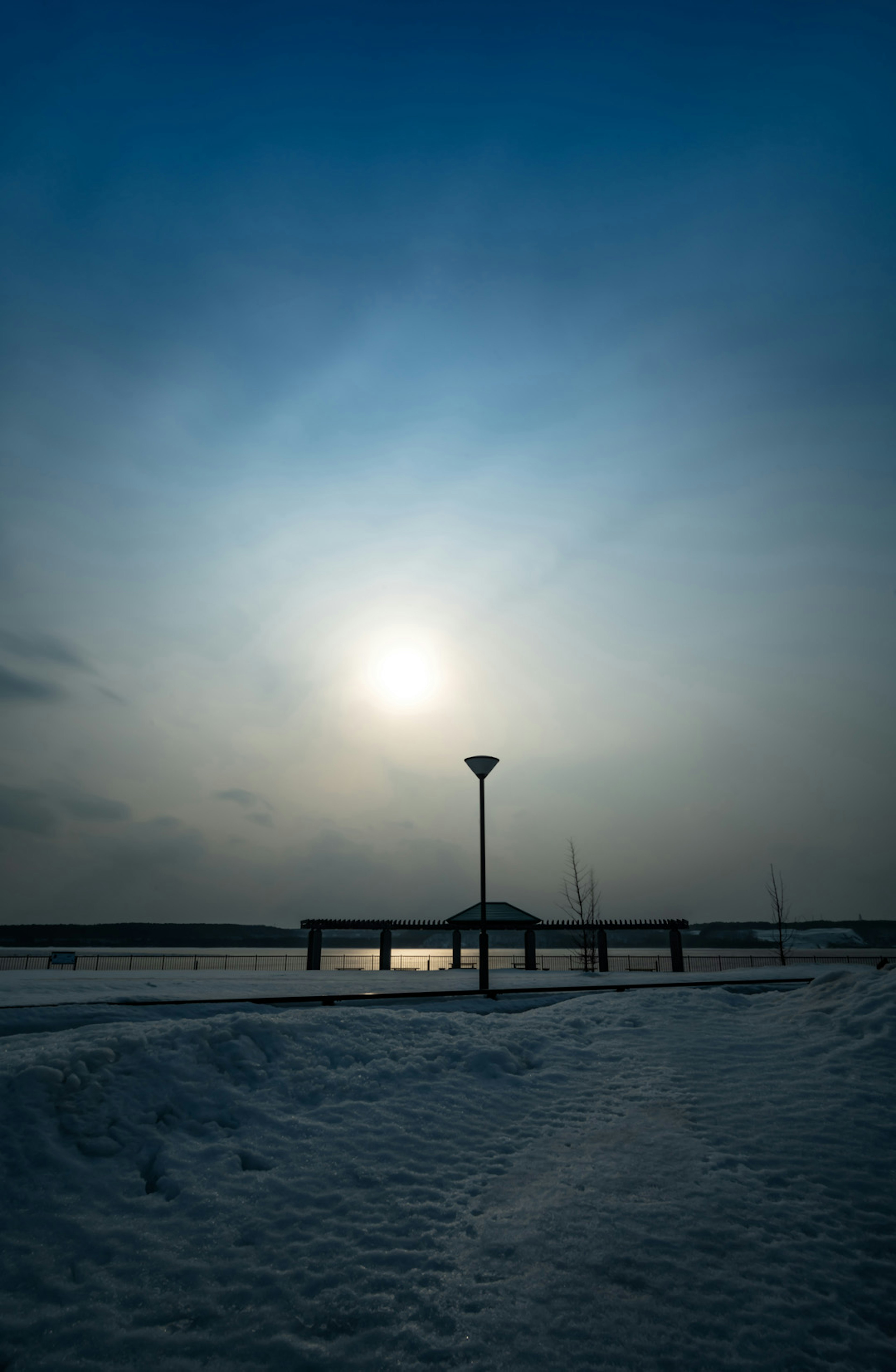 Snow-covered lakeside landscape with a setting sun and a lamp post