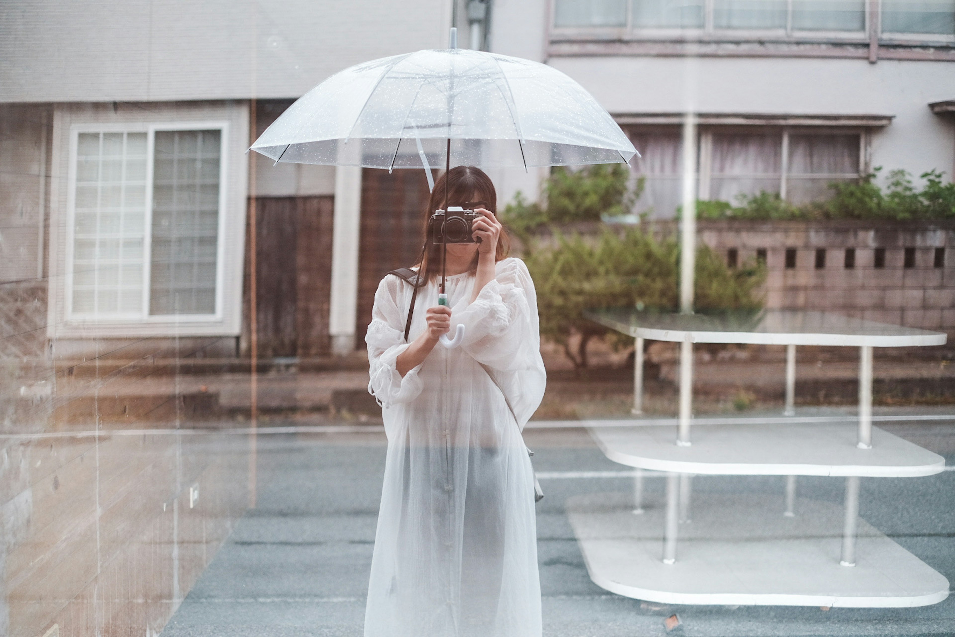 A woman in a white dress holding an umbrella and pointing a camera at the viewer seen through a window