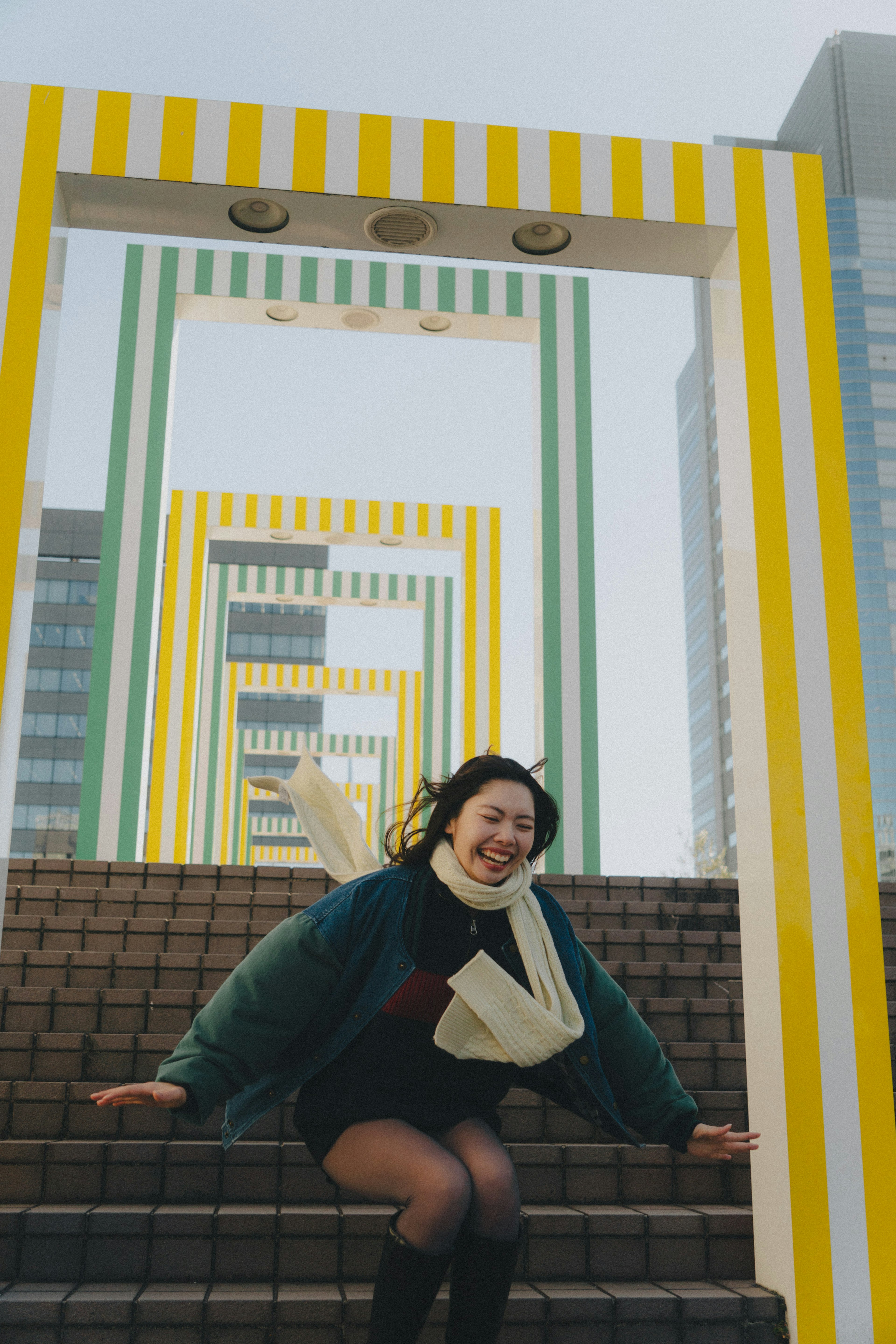 Una mujer saltando felizmente en las escaleras con arcos de rayas amarillas y verdes al fondo