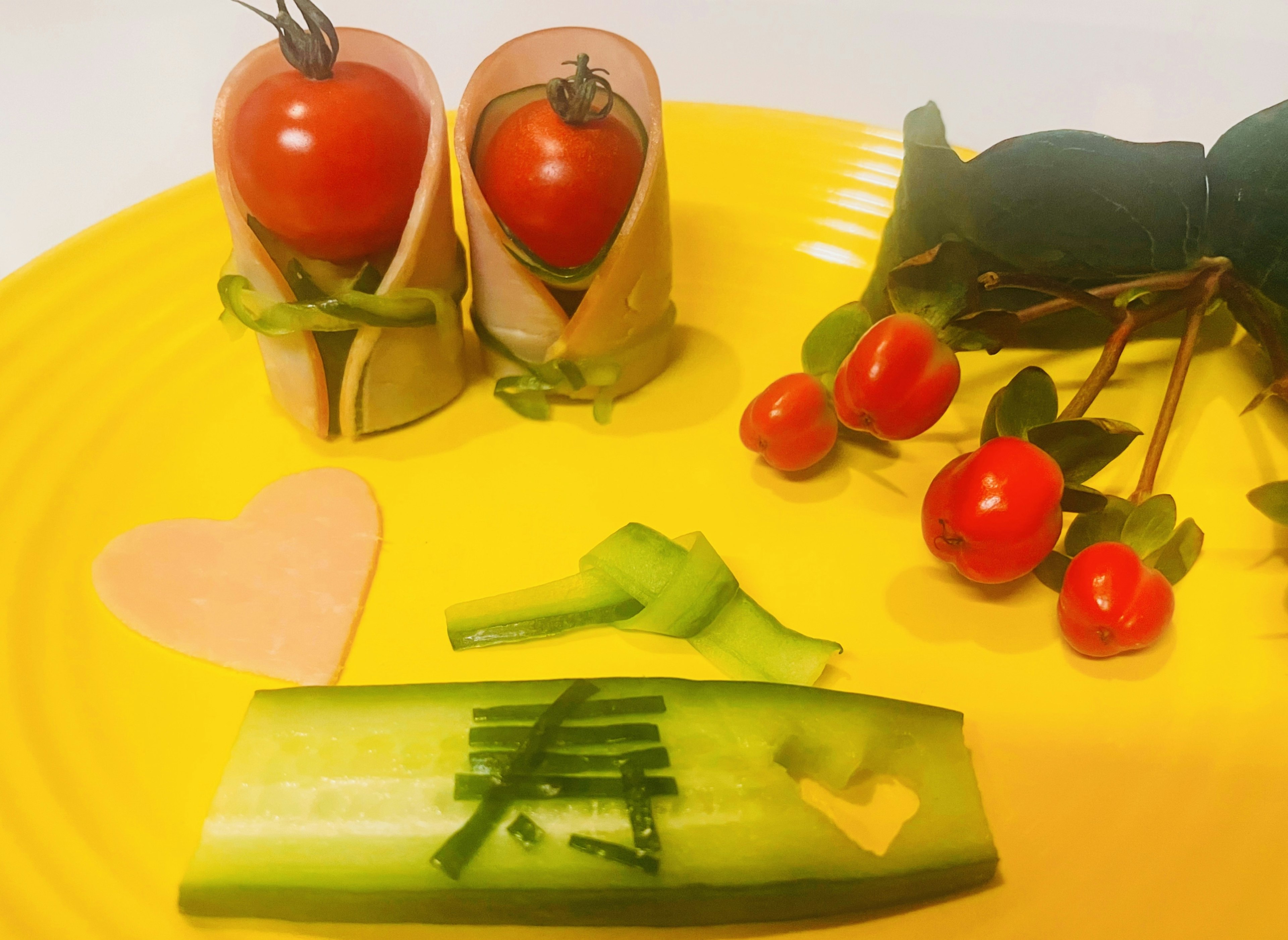 Disposizione artistica di verdure e frutta su un piatto giallo con rotoli di pomodoro e prosciutto cetriolo a forma di cuore e foglie verdi