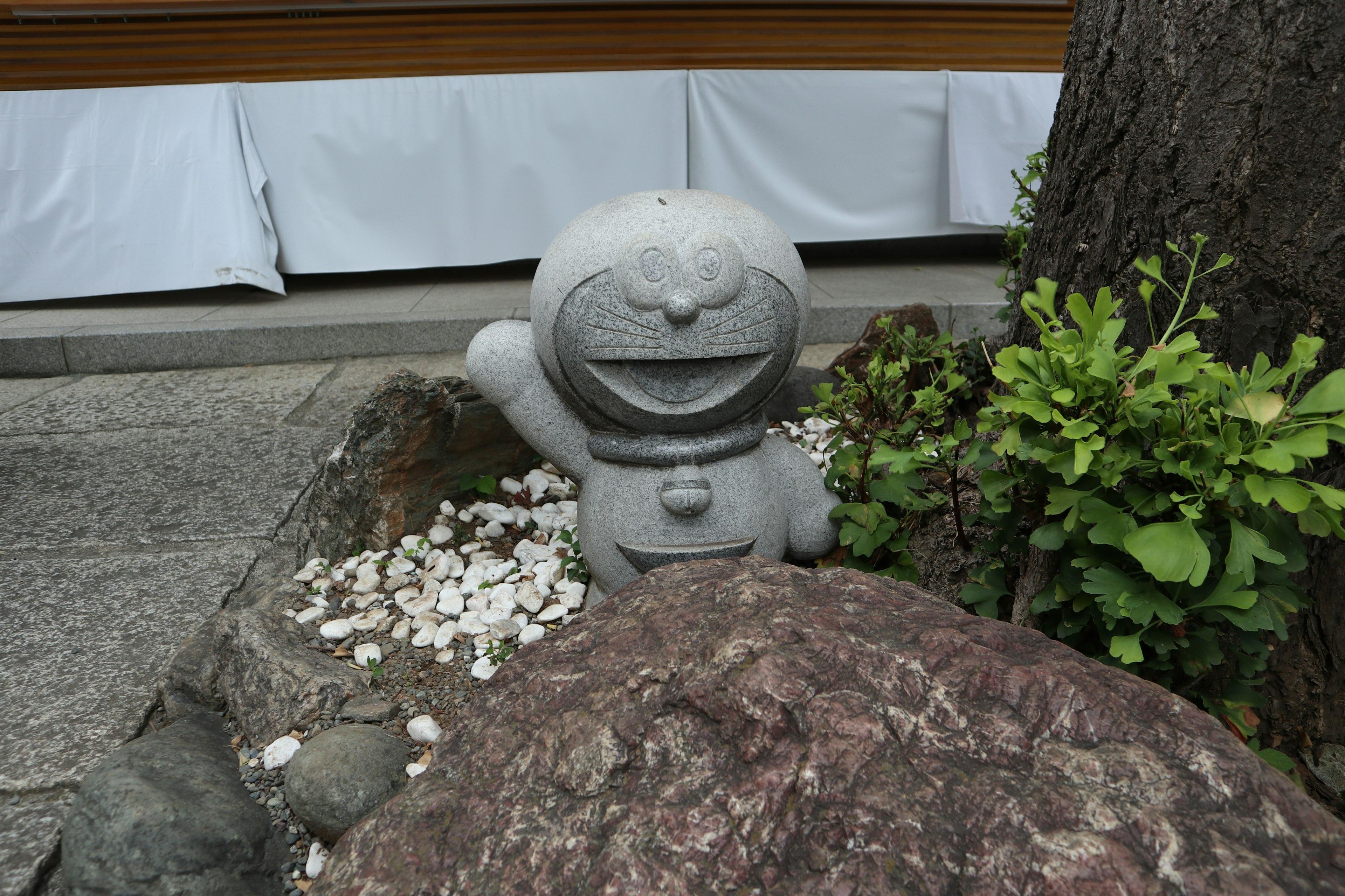 Estatua de piedra de Doraemon junto a una roca rodeada de plantas verdes y pétalos blancos